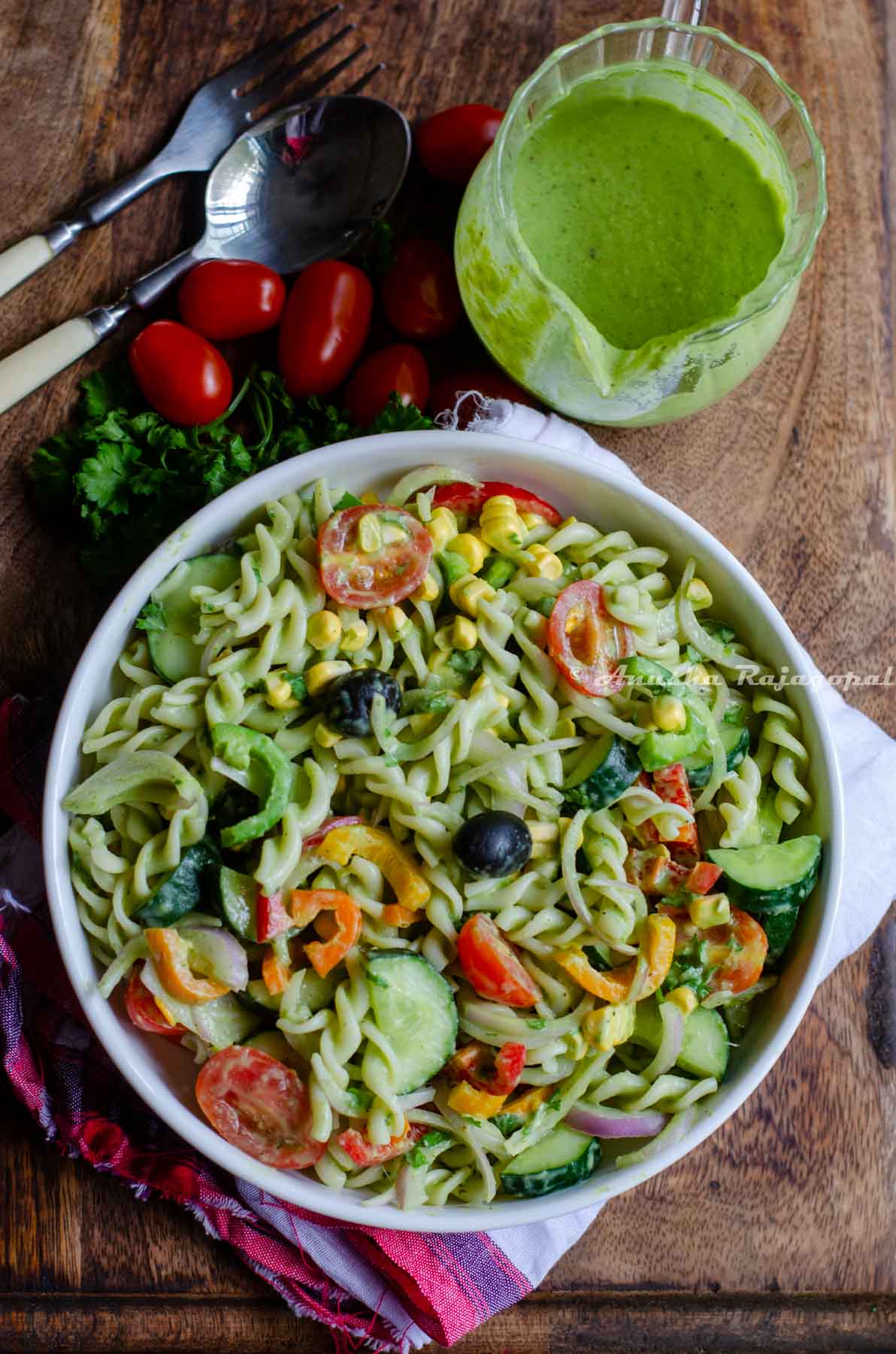 summer pasta salad served in a deep white bowl placed on a wooden board. Cutlery by the side and tomatoes and herbs around.