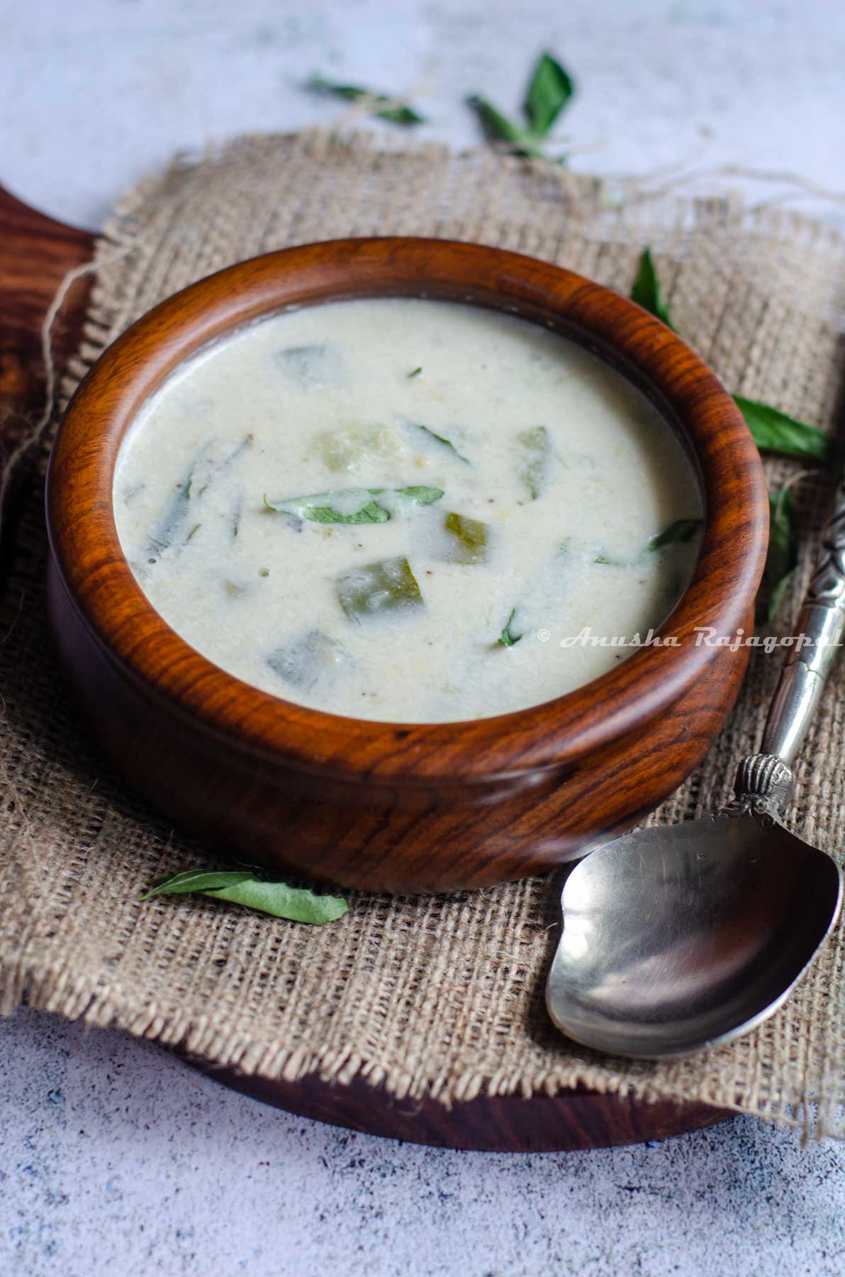 Instant pot zucchini Kootu served in a wooden bowl placed over a burlap mat. A serving spoon by the side. Curry leaves scattered at the back ground.