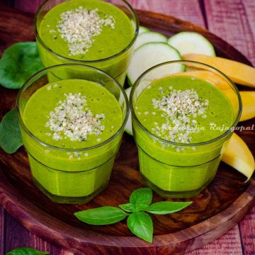 mango spinach smoothie served with hemp seed topping. Mangoes, zucchini and herbs around the smoothie glasses.