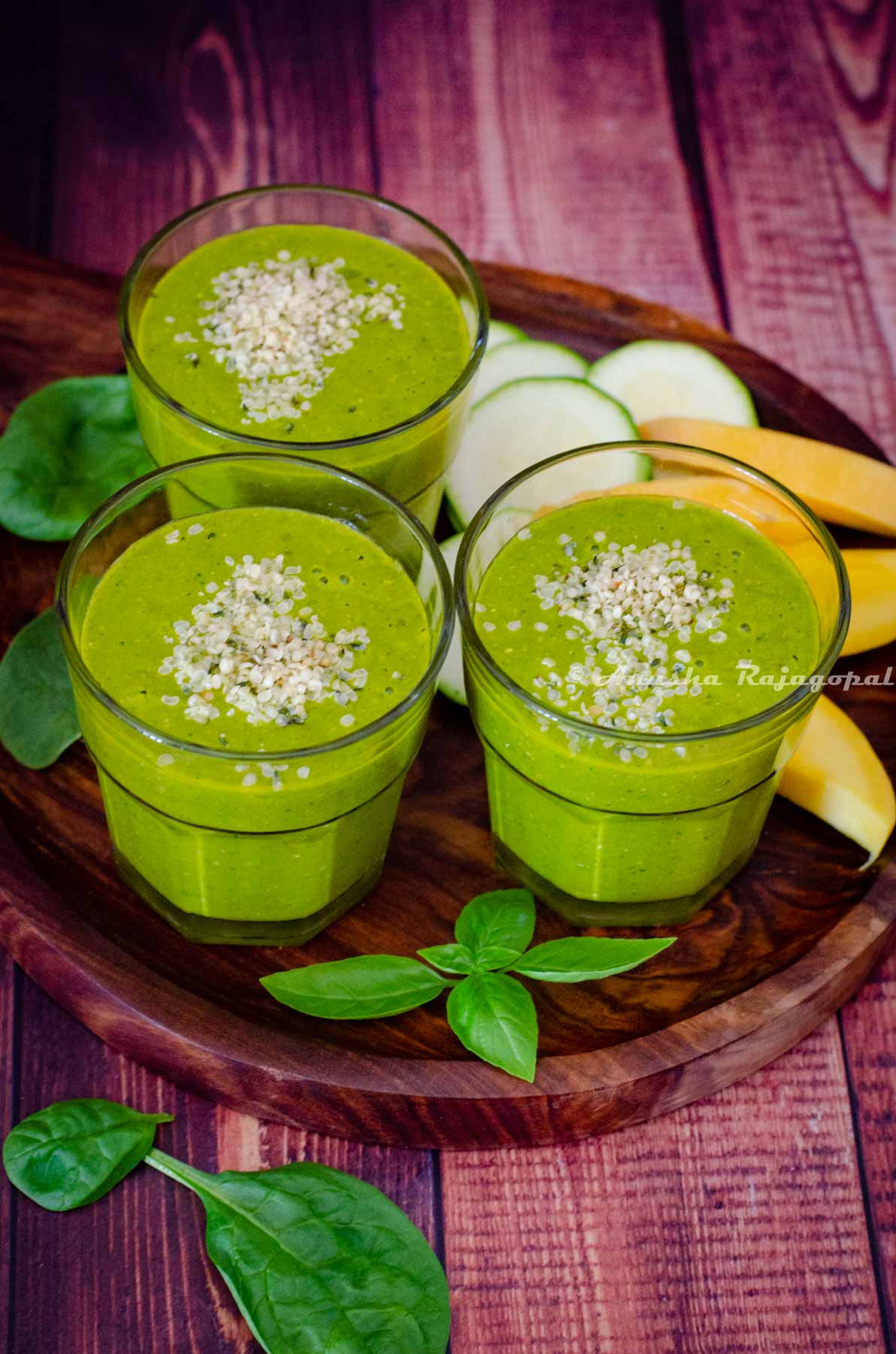 mango spinach smoothie served with hemp seed topping. Mangoes, zucchini and herbs around the smoothie glasses.