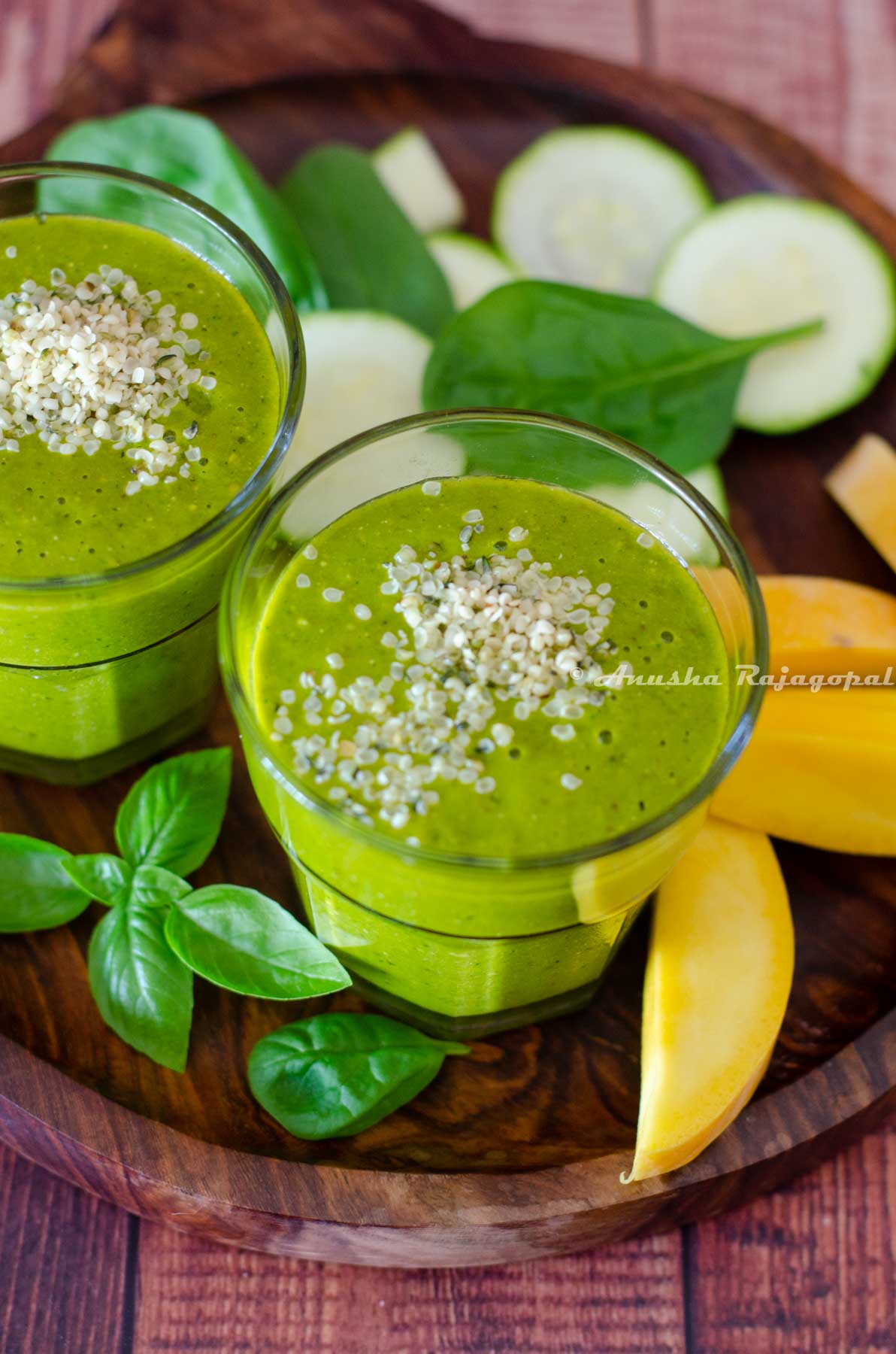 mango spinach smoothie served with hemp seed topping. Mangoes, zucchini and herbs around the smoothie glasses.