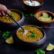 vegan persian lentil soup served in black bowls. Someone trying to scoop out some with a black handled spoon. Lime wedges and yogurt in wooden bowls behind.