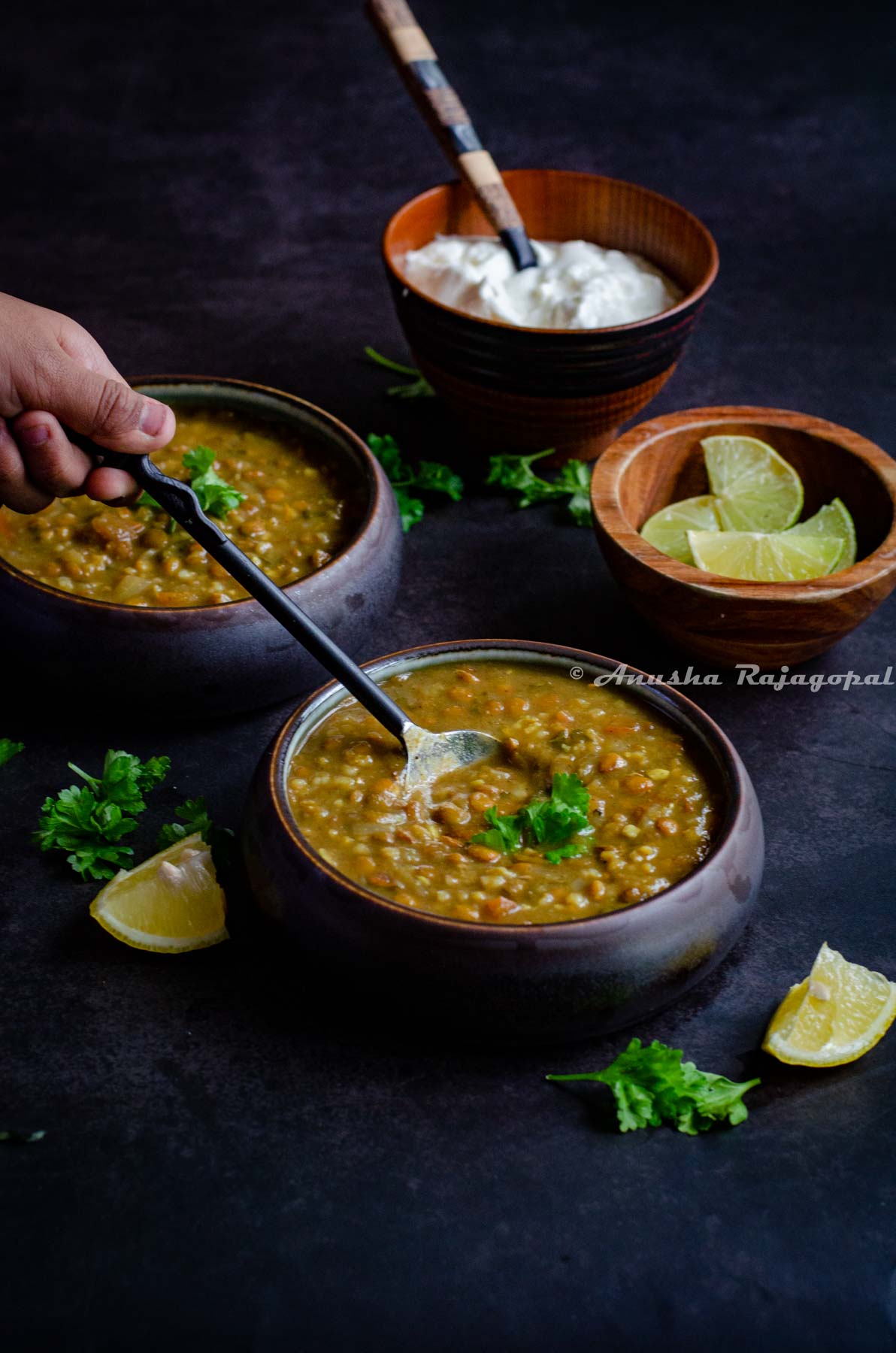 vegan persian lentil soup served in black bowls. Someone trying to scoop out some with a black handled spoon. Lime wedges and yogurt in wooden bowls behind.