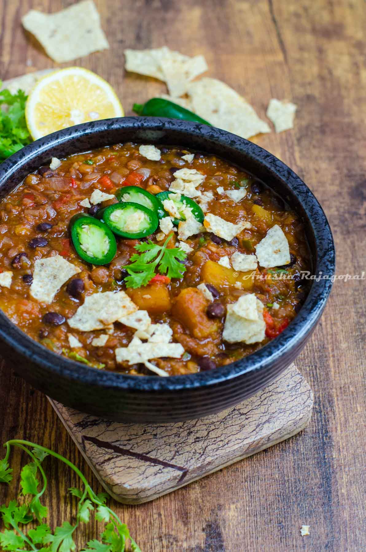 Vegan chili with butternut and lentils in a black bowl placed on a wooden cheese board. Crushed tortillas scattered by the side.