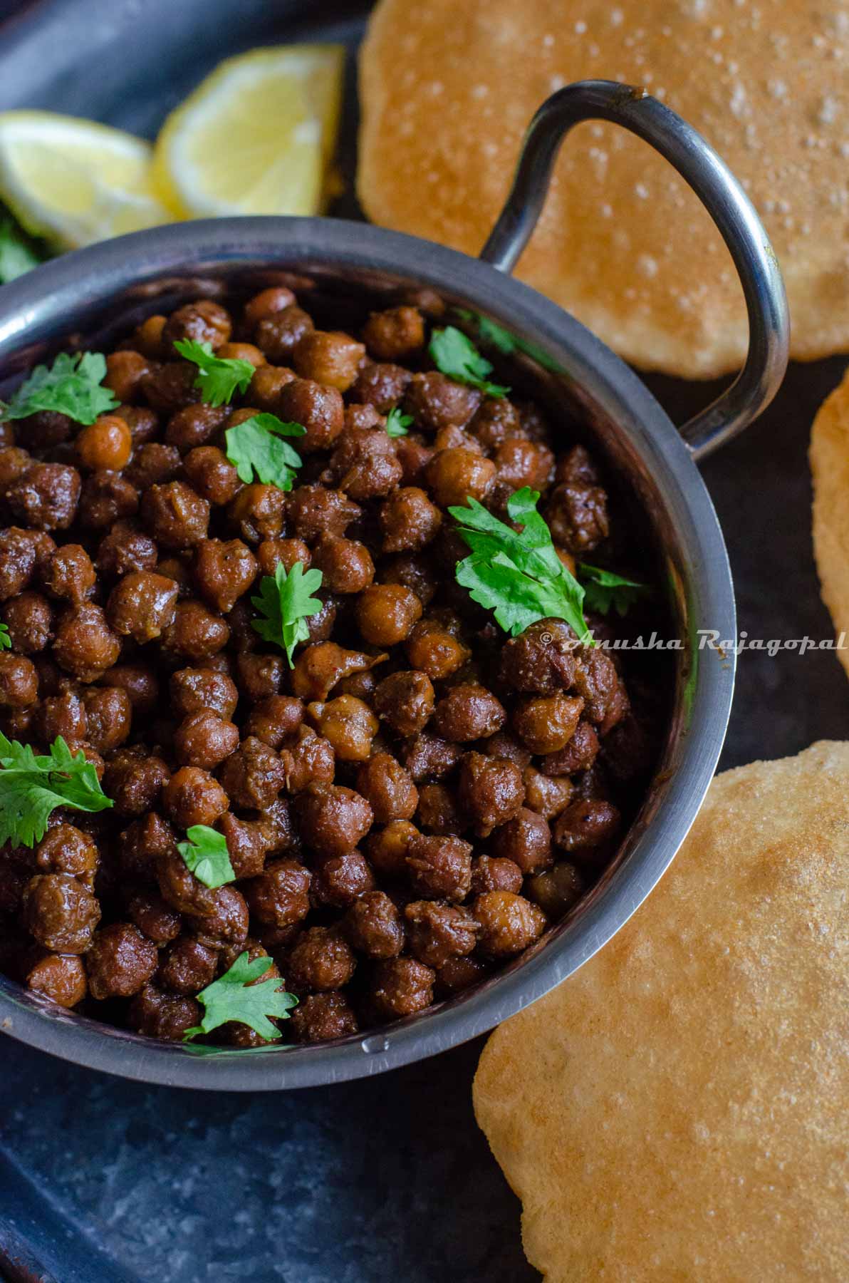 Kala chana made in Instant pot served in a steel pan with pooris placed on a black platter