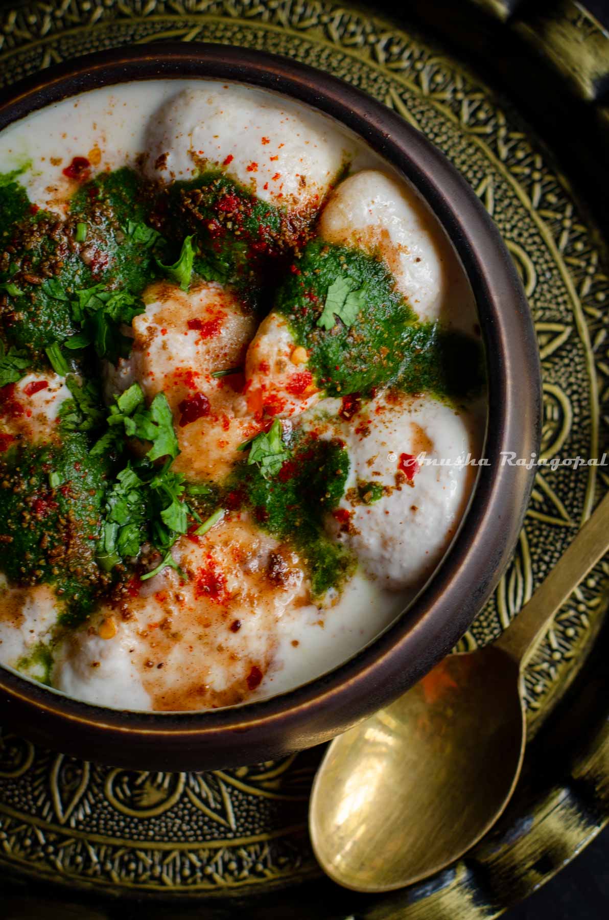 soft dahi bhalle served with chutneys and spices in a black bowl. Bowl placed over an antique charger plate with a brass spoon by the side.