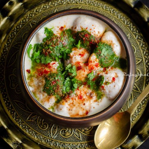 soft dahi bhalle served with chutneys and spices in a black bowl. Bowl placed over an antique charger plate with a brass spoon by the side.