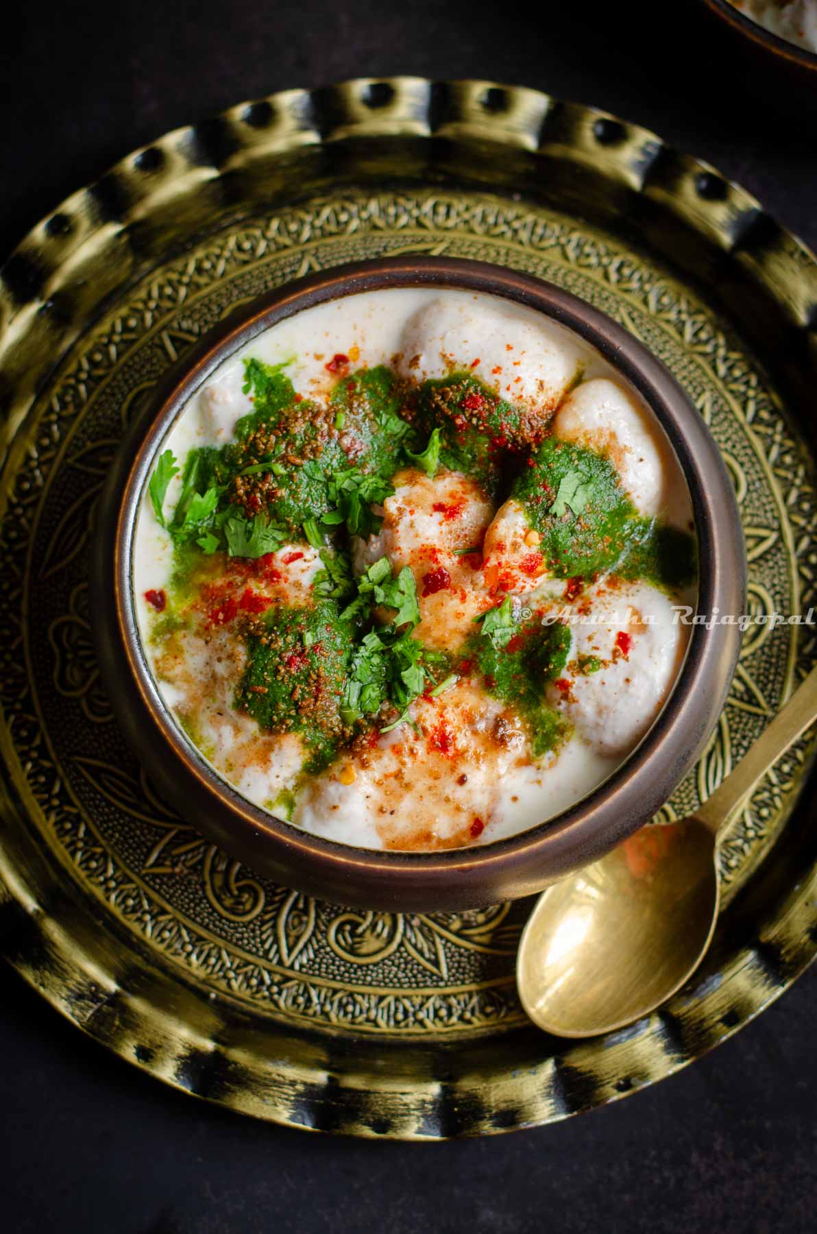soft dahi bhalle served with chutneys and spices in a black bowl. Bowl placed over an antique charger plate with a brass spoon by the side.