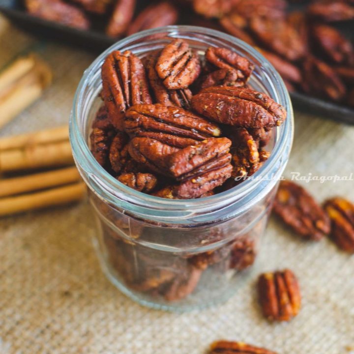 air fryer spiced pecans in a jar placed on a burlap mat. Pecans strewn around and a baking tray of pecans at the background. Cinnamon quills by the left side.
