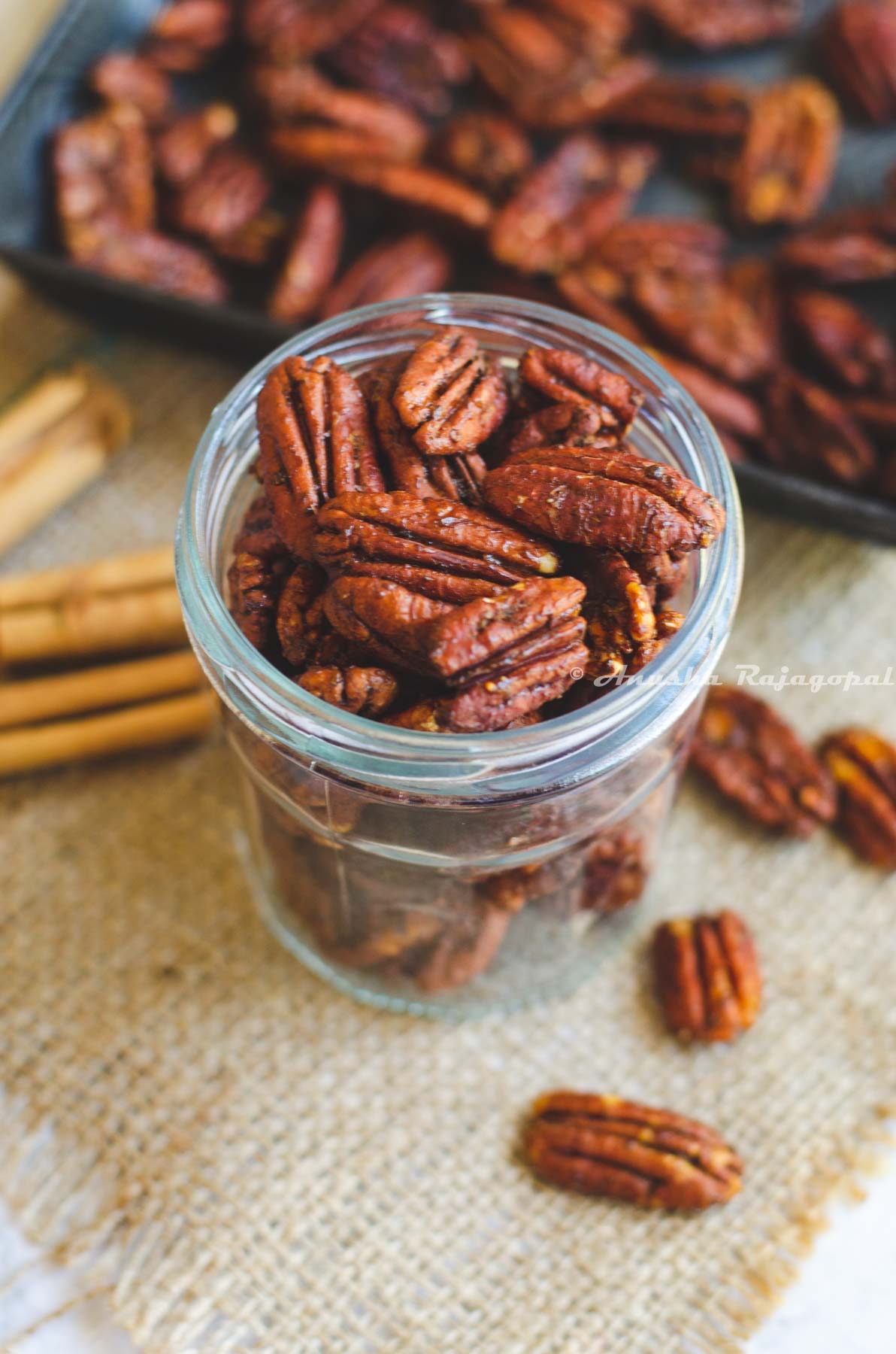 air fryer spiced pecans in a jar placed on a burlap mat. Pecans strewn around and a baking tray of pecans at the background. Cinnamon quills by the left side.