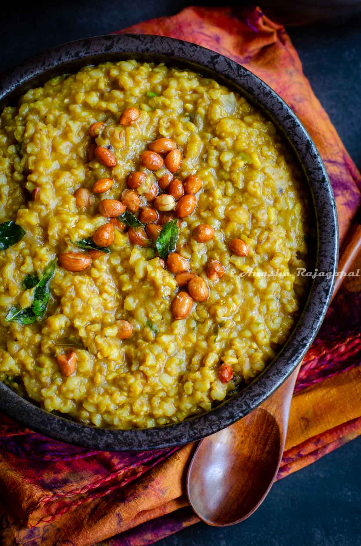 Instant pot sambar rice served in a black bowl topped with roasted peanuts. The bowl is placed over a tye-dye patterned napkin. A spoon is also by the side of the bowl.