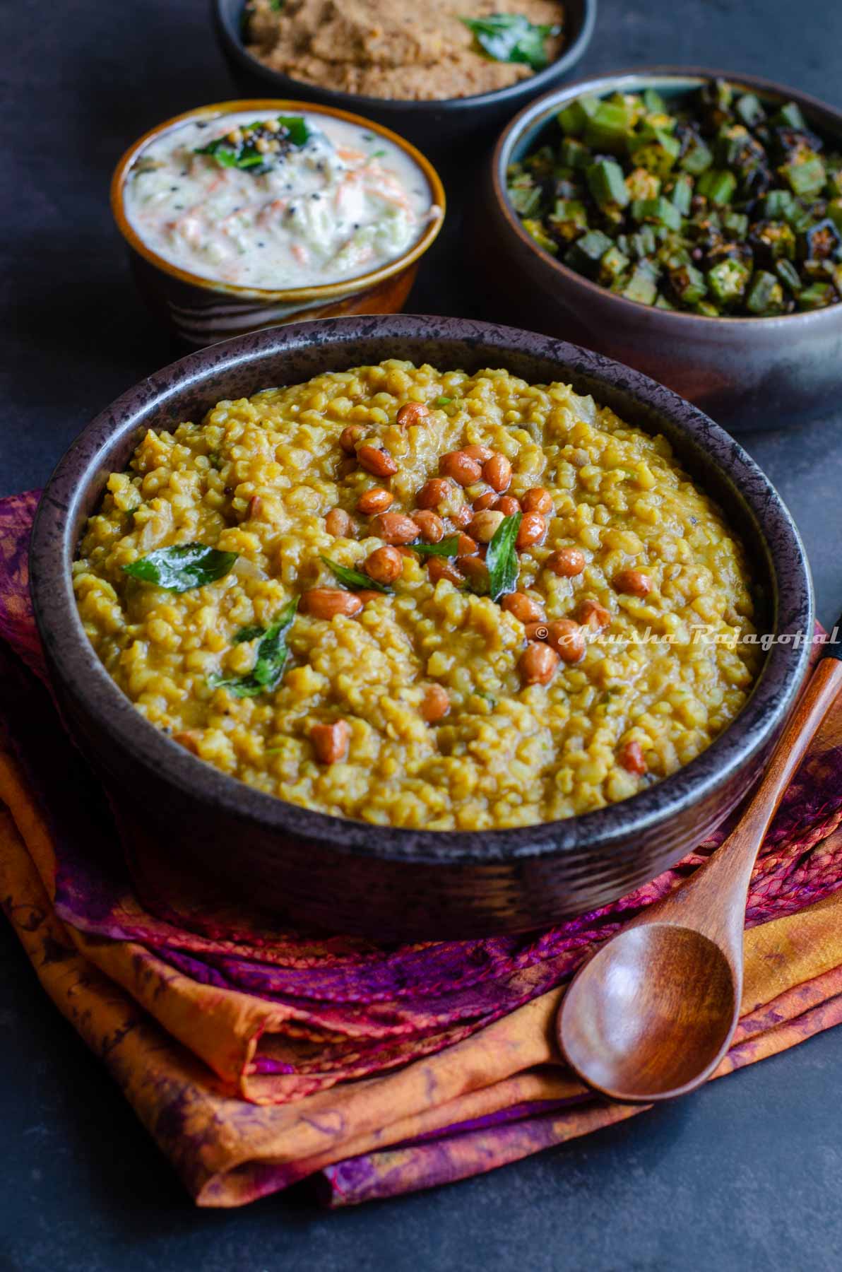 Instant pot sambar rice served in a black bowl topped with roasted peanuts. The bowl is placed over a tye-dye patterned napkin. A spoon is also by the side of the bowl.