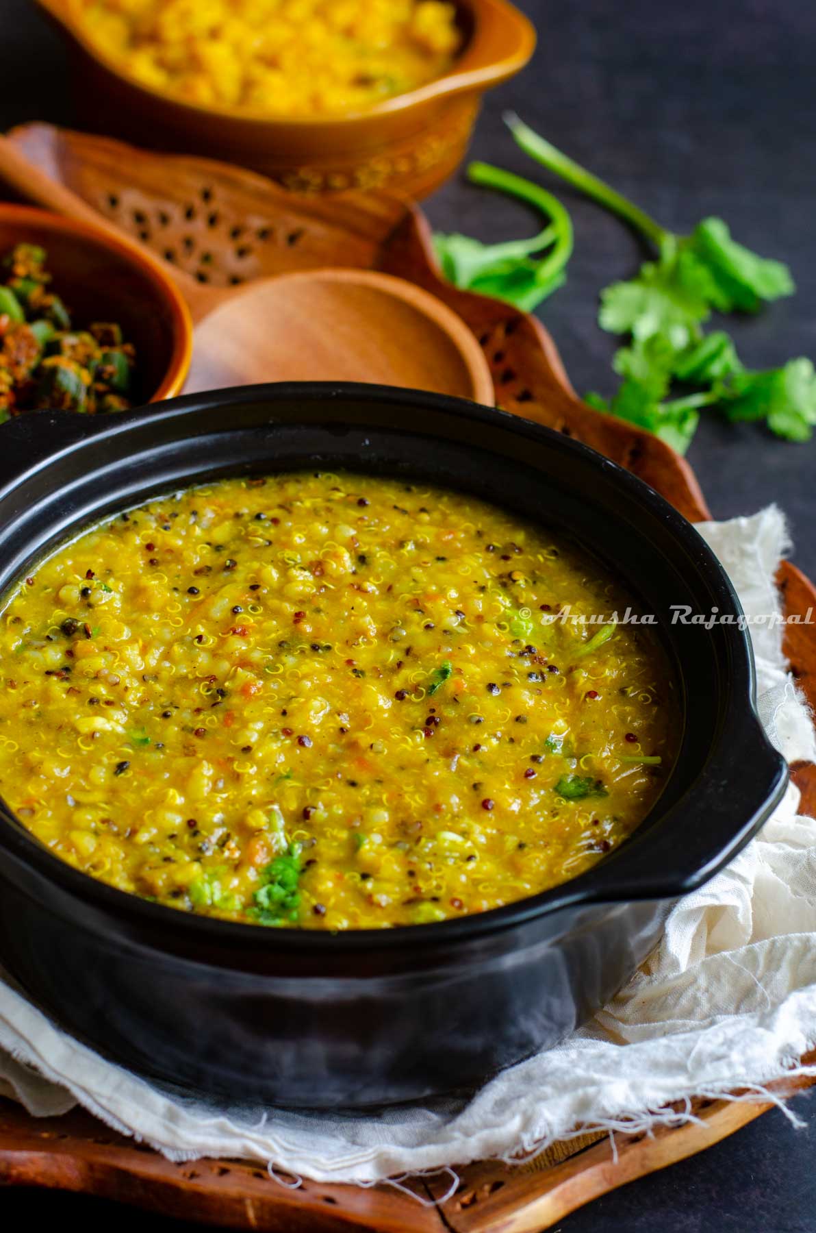 Instant pot quinoa rasam rice served in a black stew pot. Okra curry at the background. All this placed on a linen cloth over a wooden tray on a black table top. Wooden ladle and fresh coriander placed for aesthetics.