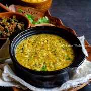 Instant pot quinoa rasam rice served in a black stew pot. Okra curry at the background. All this placed on a linen cloth over a wooden tray on a black table top. Wooden ladle and fresh coriander placed for aesthetics.