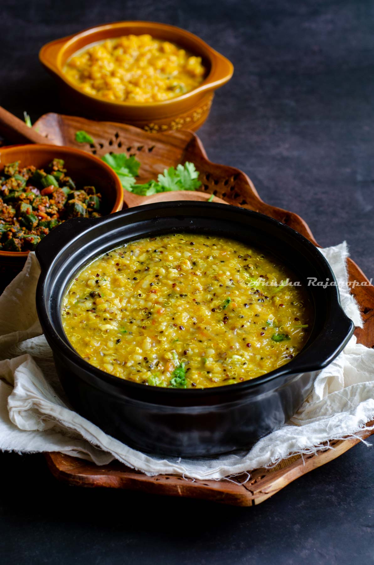 Instant pot quinoa rasam rice served in a black stew pot. Okra curry at the background. All this placed on a linen cloth over a wooden tray on a black table top. Wooden ladle and fresh coriander placed for aesthetics.