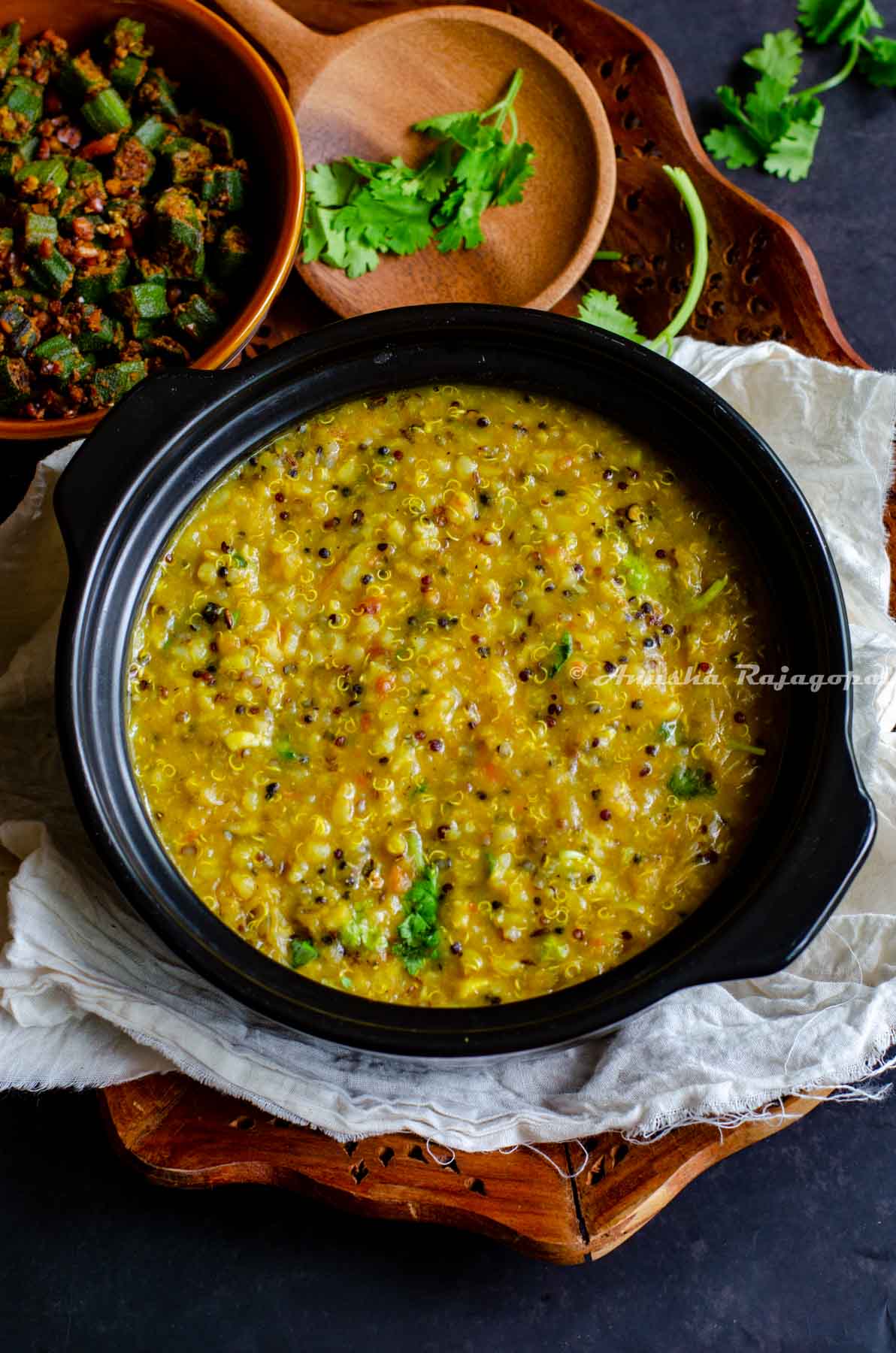 Instant pot quinoa rasam rice served in a black stew pot. Okra curry at the background. All this placed on a linen cloth over a wooden tray on a black table top. Wooden ladle and fresh coriander placed for aesthetics.