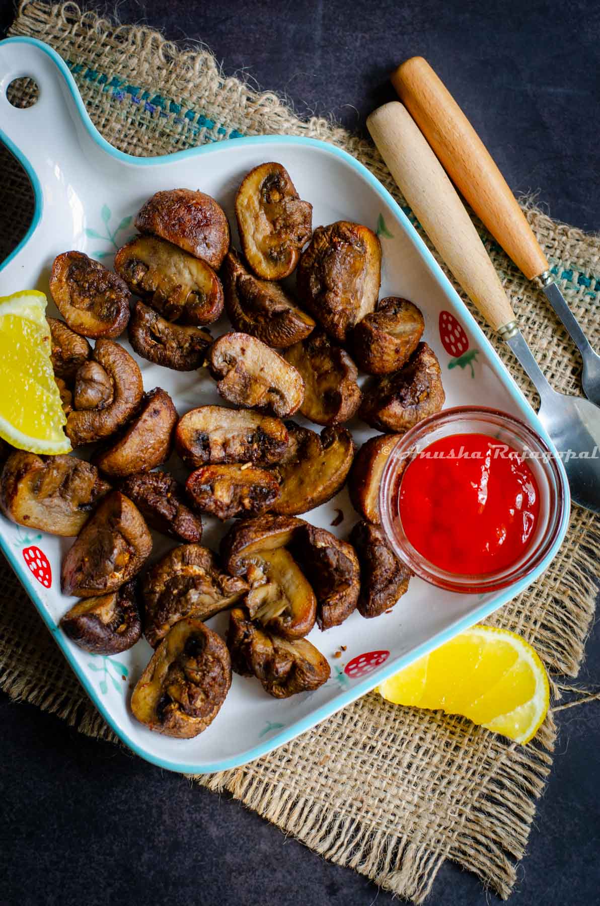 Air fried mushrooms and a small glass dip bowl with sriracha sauce served in a  blue rimmed white square platter. Lemon wedge on the mushrooms. Platter placed on a burlap mat. Spoon and fork with wooden handle by the side. 