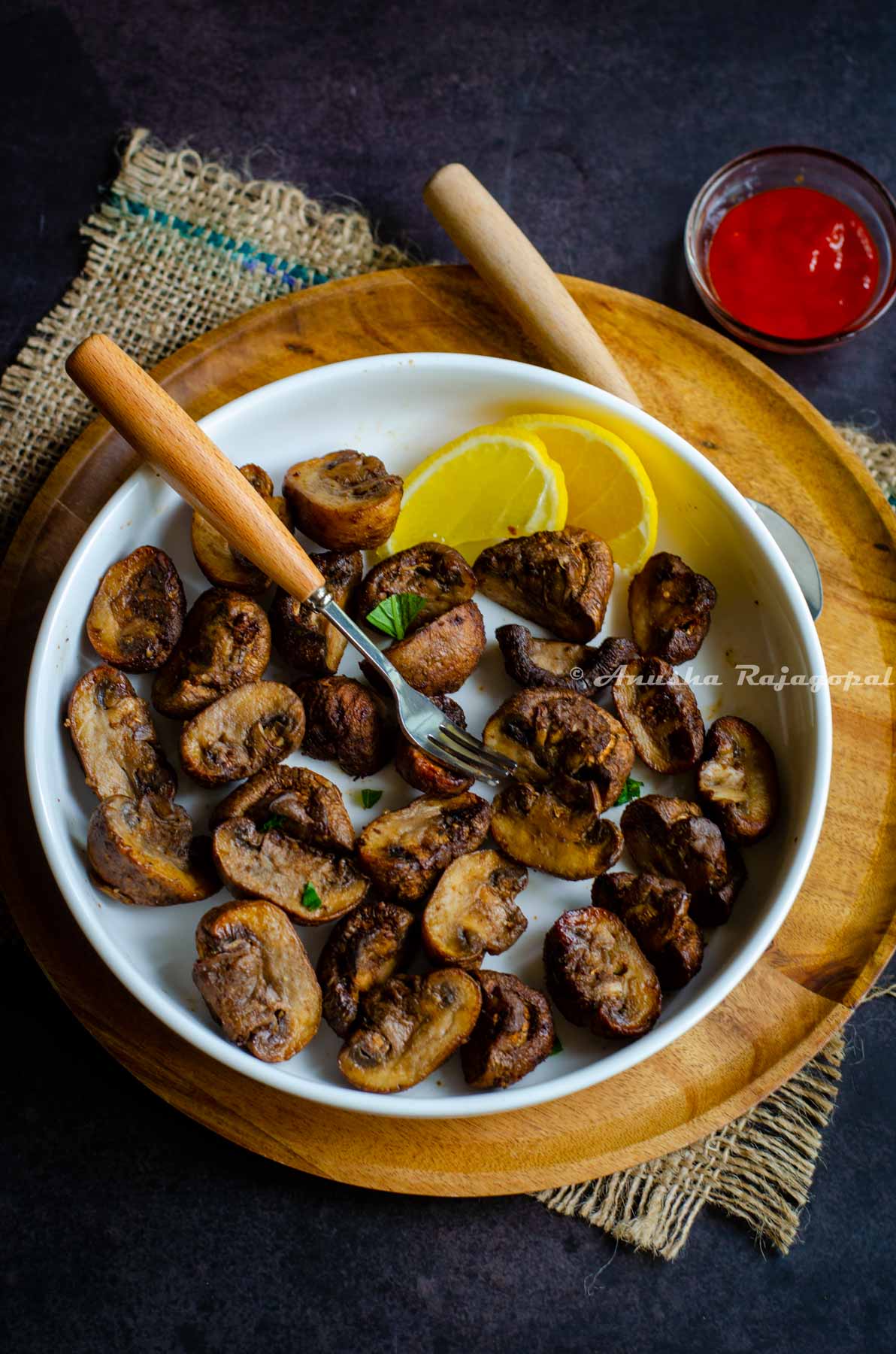 air fryer mushrooms served in a deep white platter placed on a wooden plate. Wooden plate placed over a burlap mat. Lemon wedges and fork on the mushrooms. A small dip bowl with sriracha by the side.