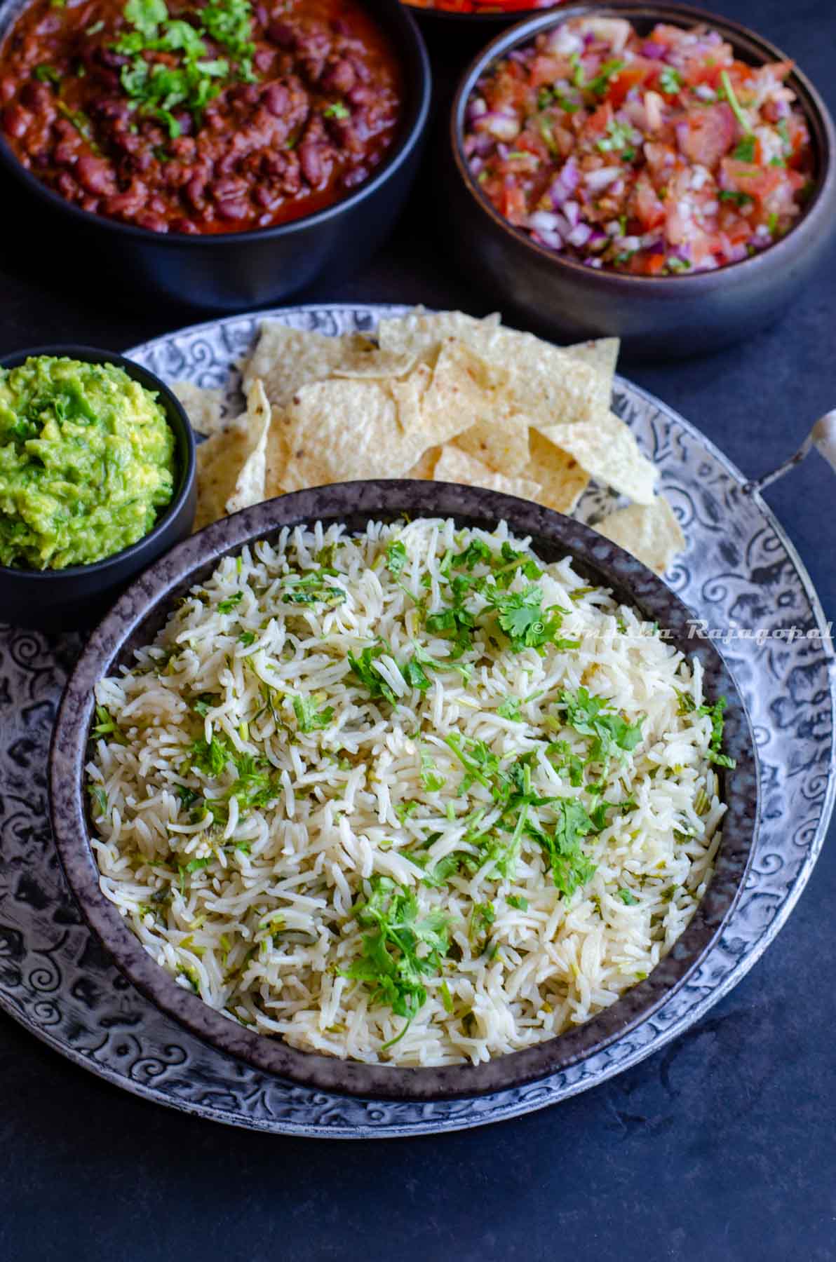 Mexican style cilantro lime rice served in a black dish with guacamole, tortilla chips and other condiments at the background.