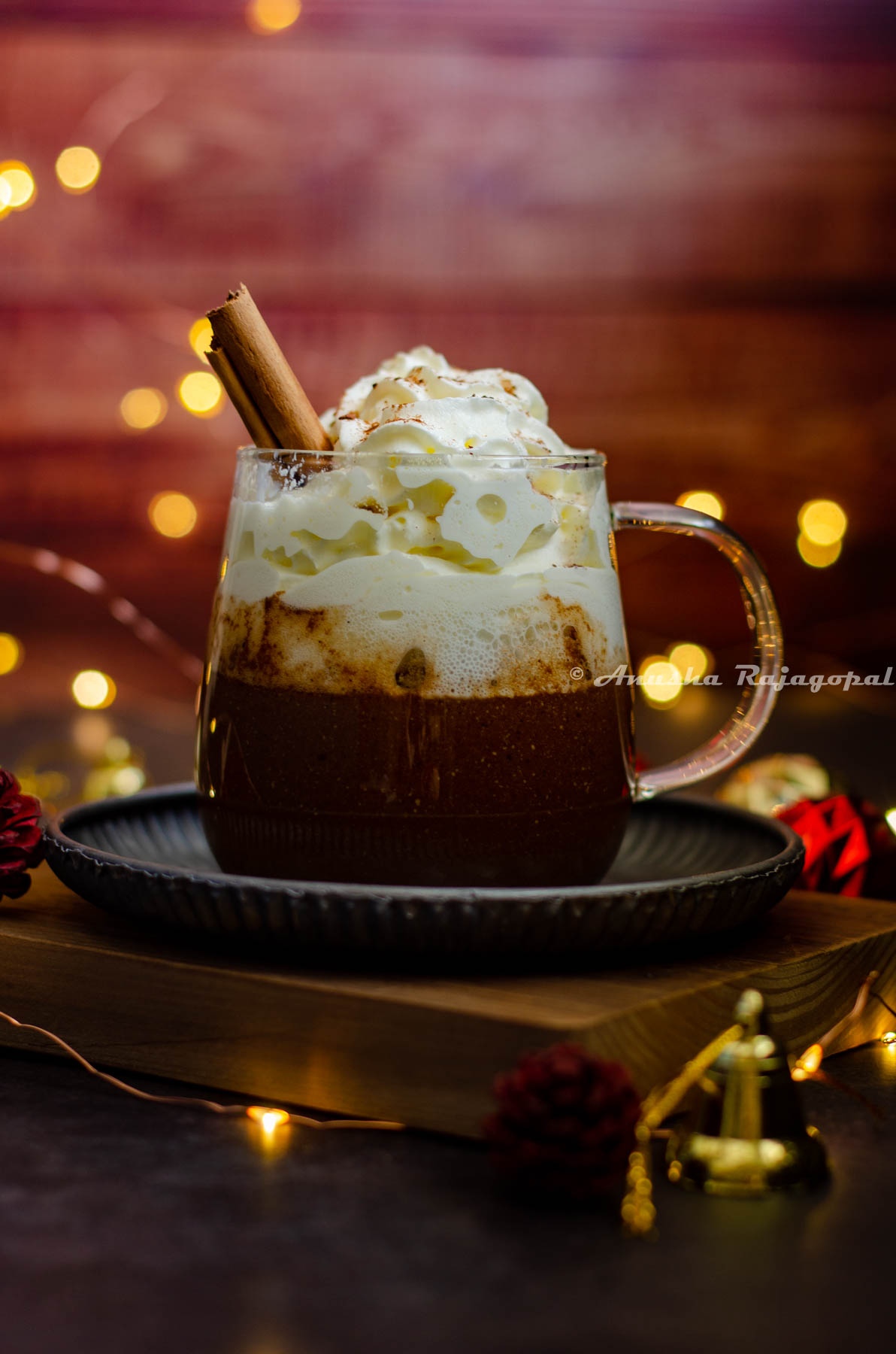 gingerbread flavored hot chocolate served in a glass mug with whipped cream topping and a sprinkle of ground cinnamon. Fairy lights and christmas tree decor surround the mug. Mug sits on a metal tray placed on a wooden board.