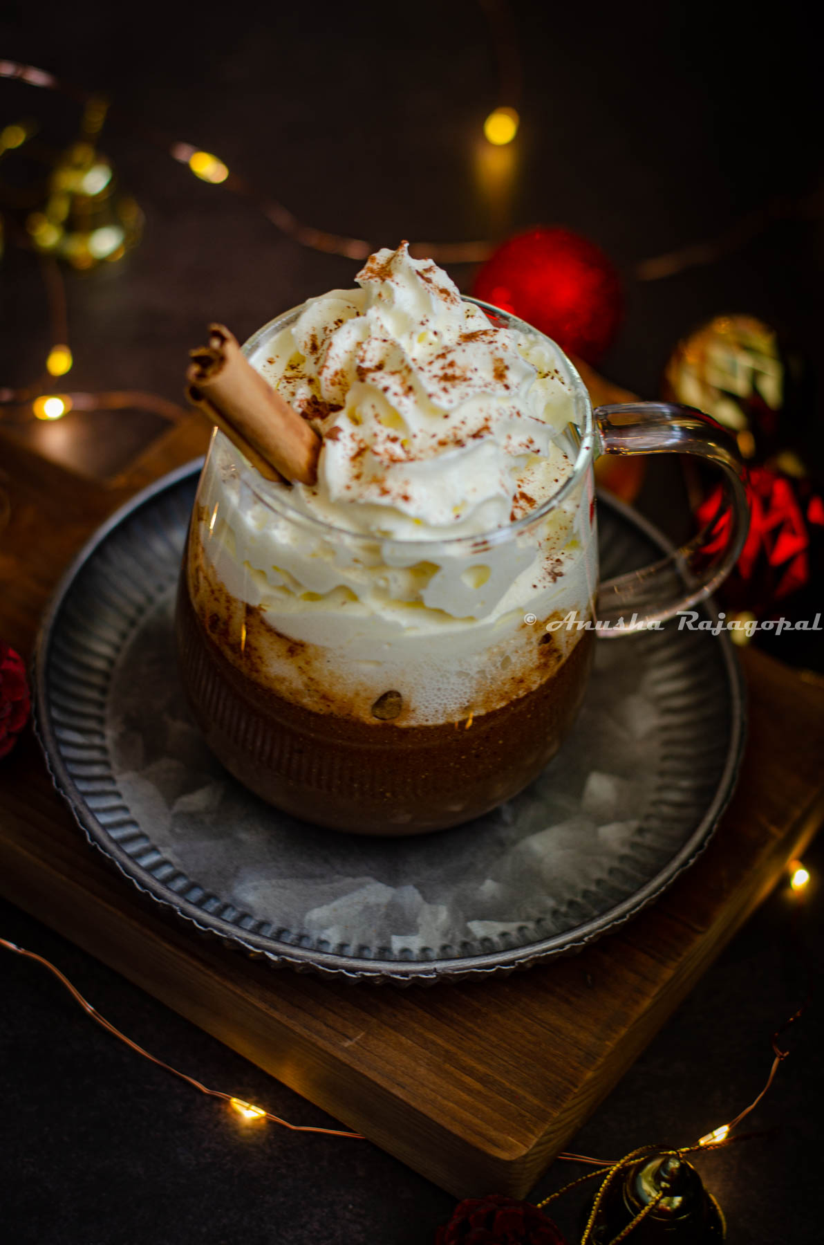 gingerbread flavored hot chocolate served in a glass mug with whipped cream topping and a sprinkle of ground cinnamon. Fairy lights and christmas tree decor surround the mug. Mug sits on a metal tray placed on a wooden board.