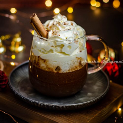 gingerbread flavored hot chocolate served in a glass mug with whipped cream topping and a sprinkle of ground cinnamon. Fairy lights and christmas tree decor surround the mug. Mug sits on a metal tray placed on a wooden board.