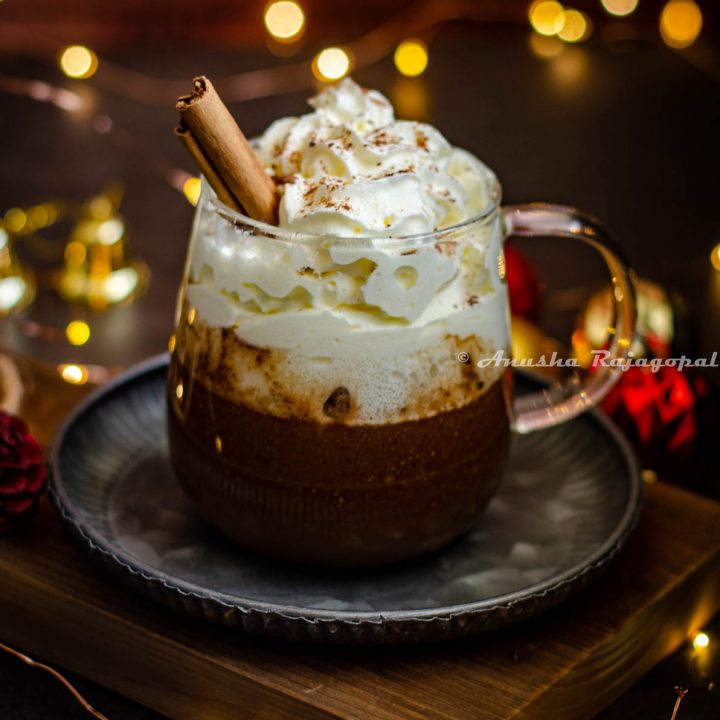 gingerbread flavored hot chocolate served in a glass mug with whipped cream topping and a sprinkle of ground cinnamon. Fairy lights and christmas tree decor surround the mug. Mug sits on a metal tray placed on a wooden board.