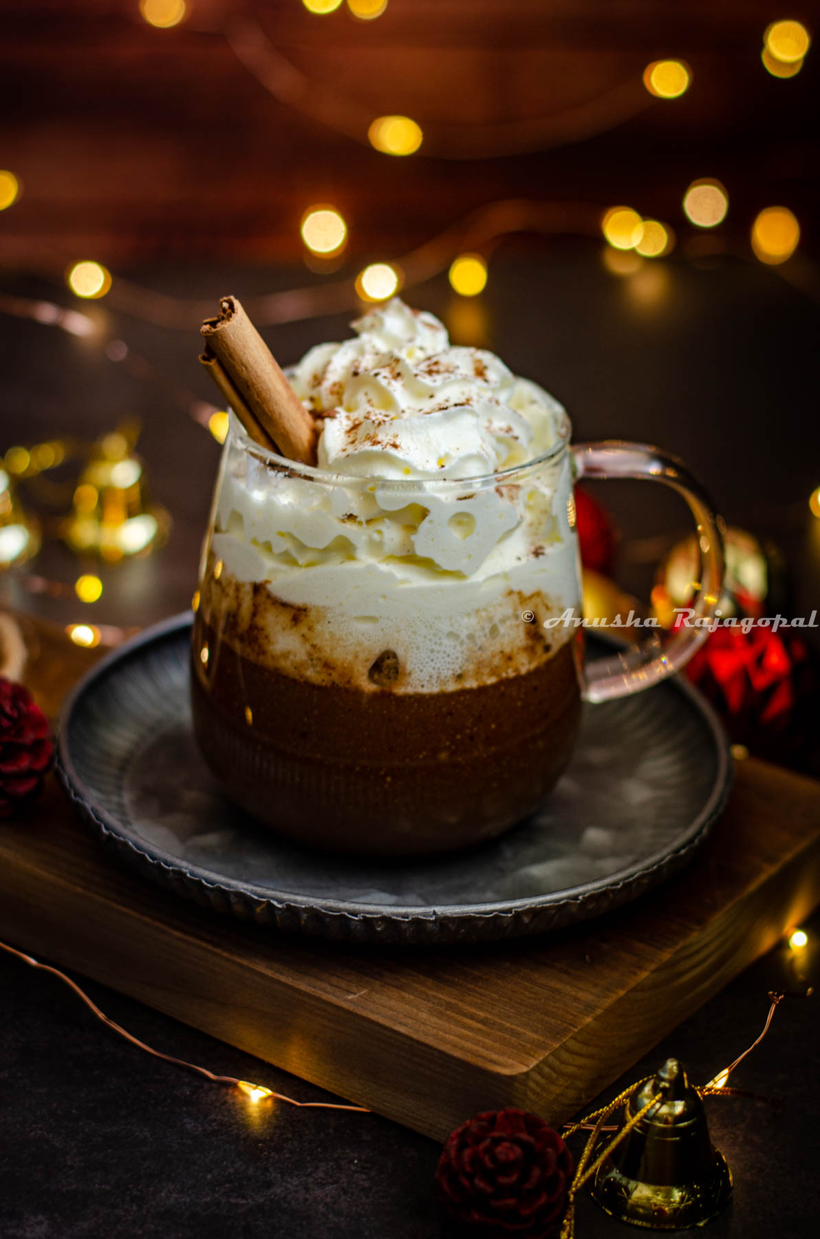 gingerbread flavored hot chocolate served in a glass mug with whipped cream topping and a sprinkle of ground cinnamon. Fairy lights and christmas tree decor surround the mug. Mug sits on a metal tray placed on a wooden board.