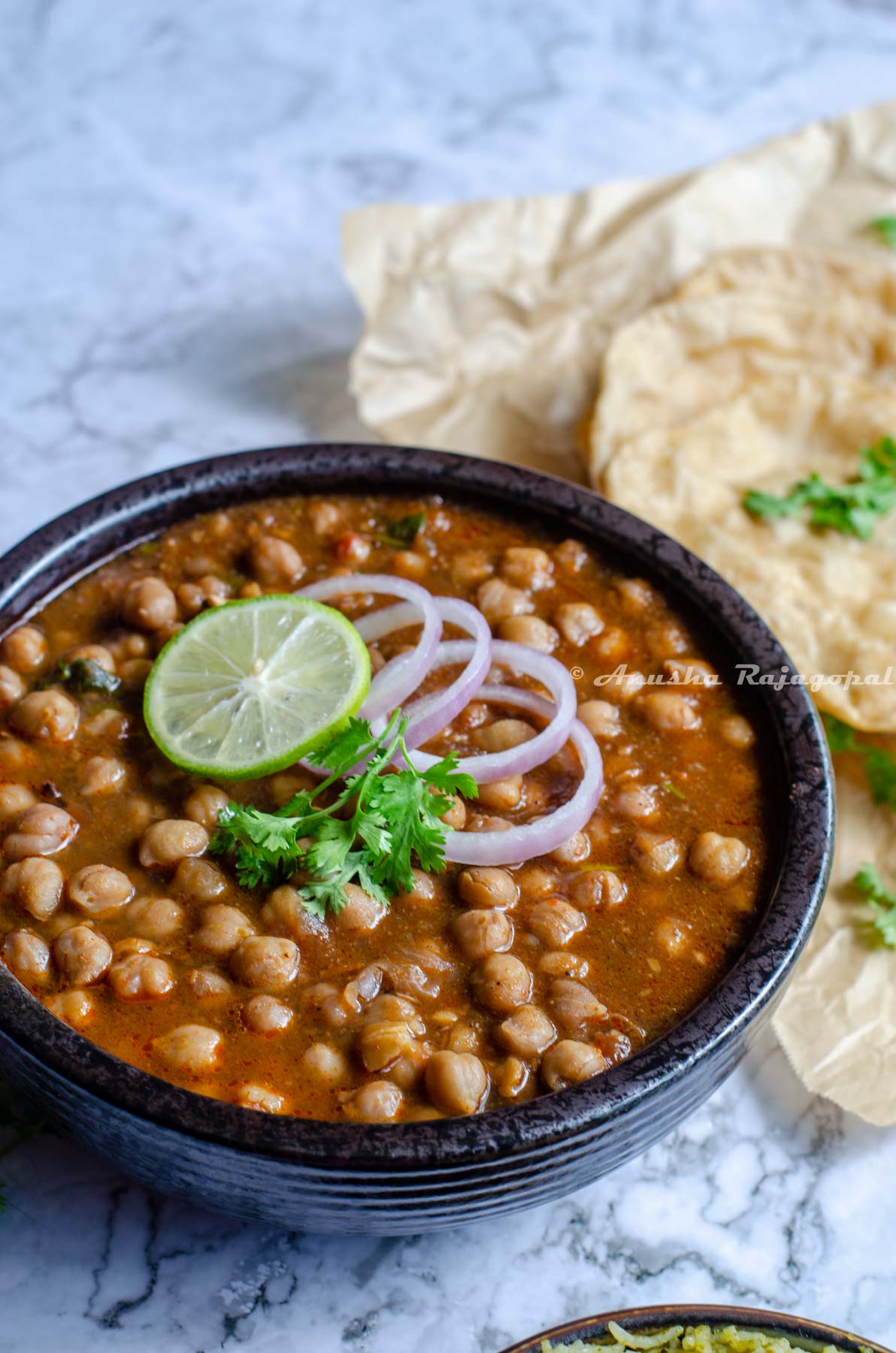 Instant pot channa masala topped with onion rings, a slice of lemon and cilantro leaves in a black bowl. The bowl is placed on a marbled surface. Pooris and at the back ground. Cilantro strewn all over.