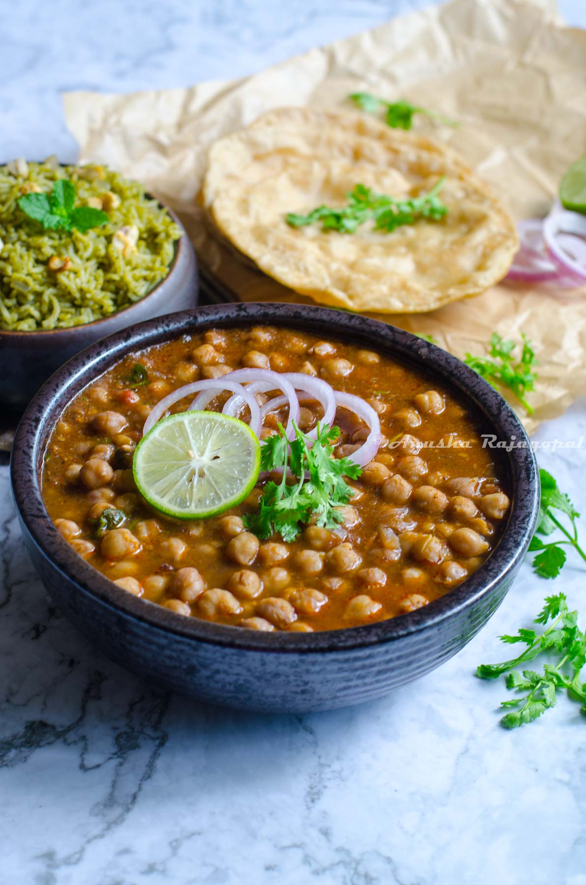 Instant pot channa masala topped with onion rings, a slice of lemon and cilantro leaves in a black bowl. The bowl is placed on a marbled surface. Pooris and a mint pilaf at the back ground. Cilantro strewn all over.