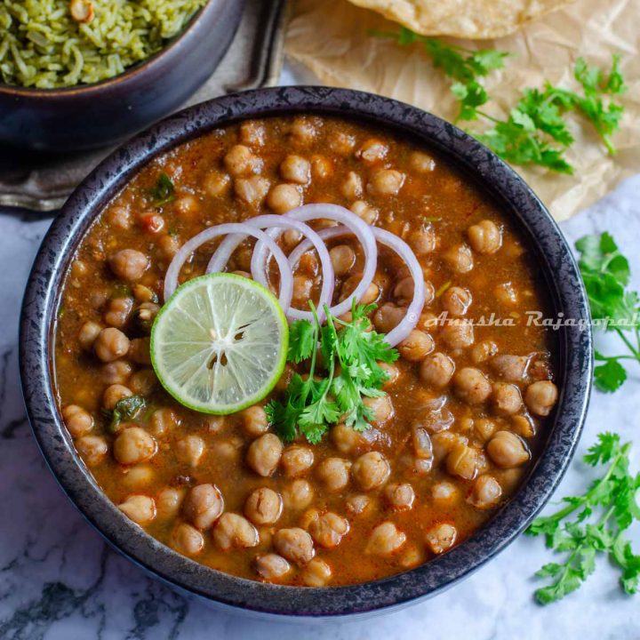 Instant pot channa masala topped with onion rings, a slice of lemon and cilantro leaves in a black bowl. The bowl is placed on a marbled surface. Pooris and a mint pilaf at the back ground. Cilantro strewn all over.