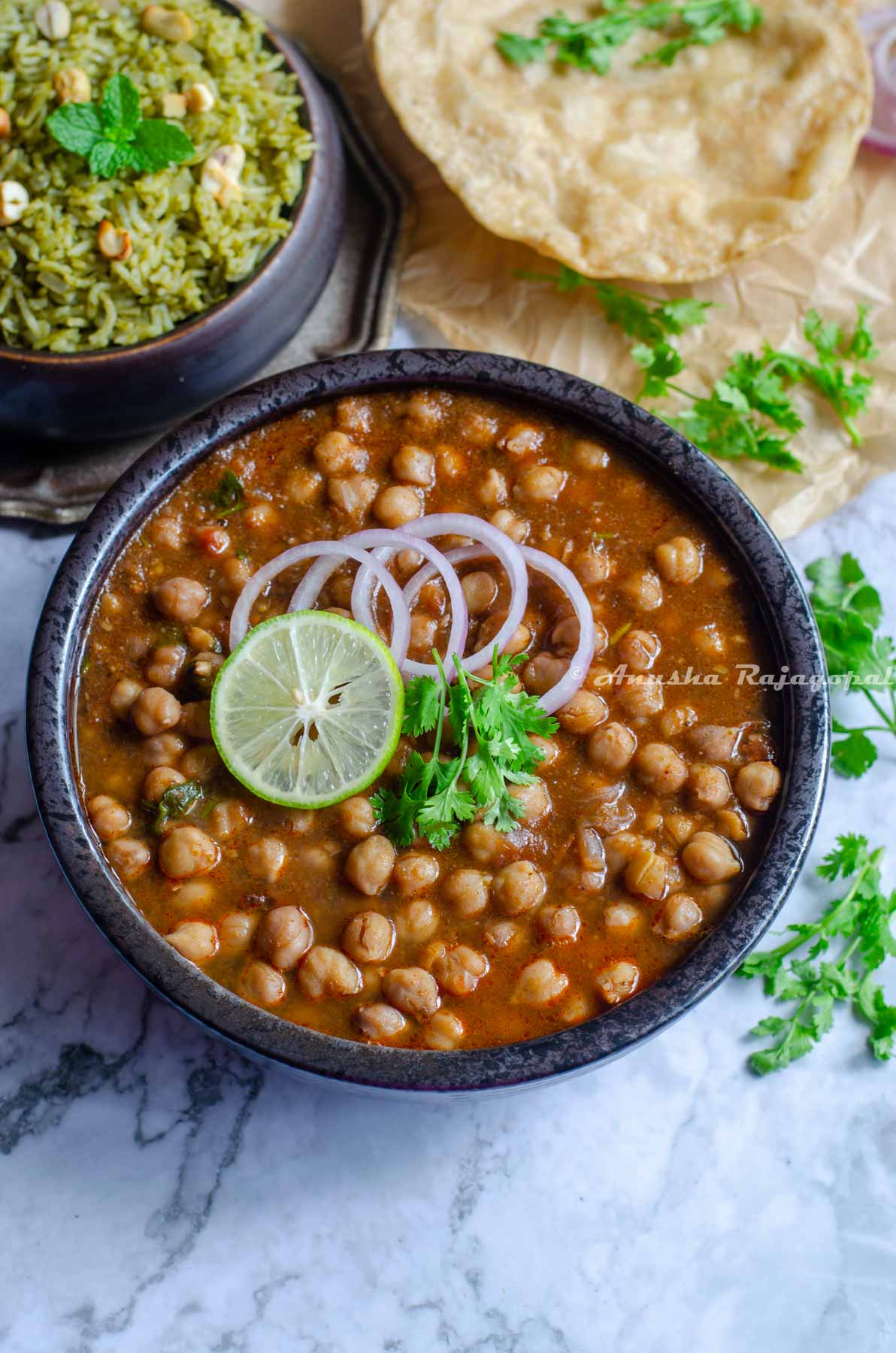Instant pot channa masala topped with onion rings, a slice of lemon and cilantro leaves in a black bowl. The bowl is placed on a marbled surface. Pooris and a mint pilaf at the back ground. Cilantro strewn all over.