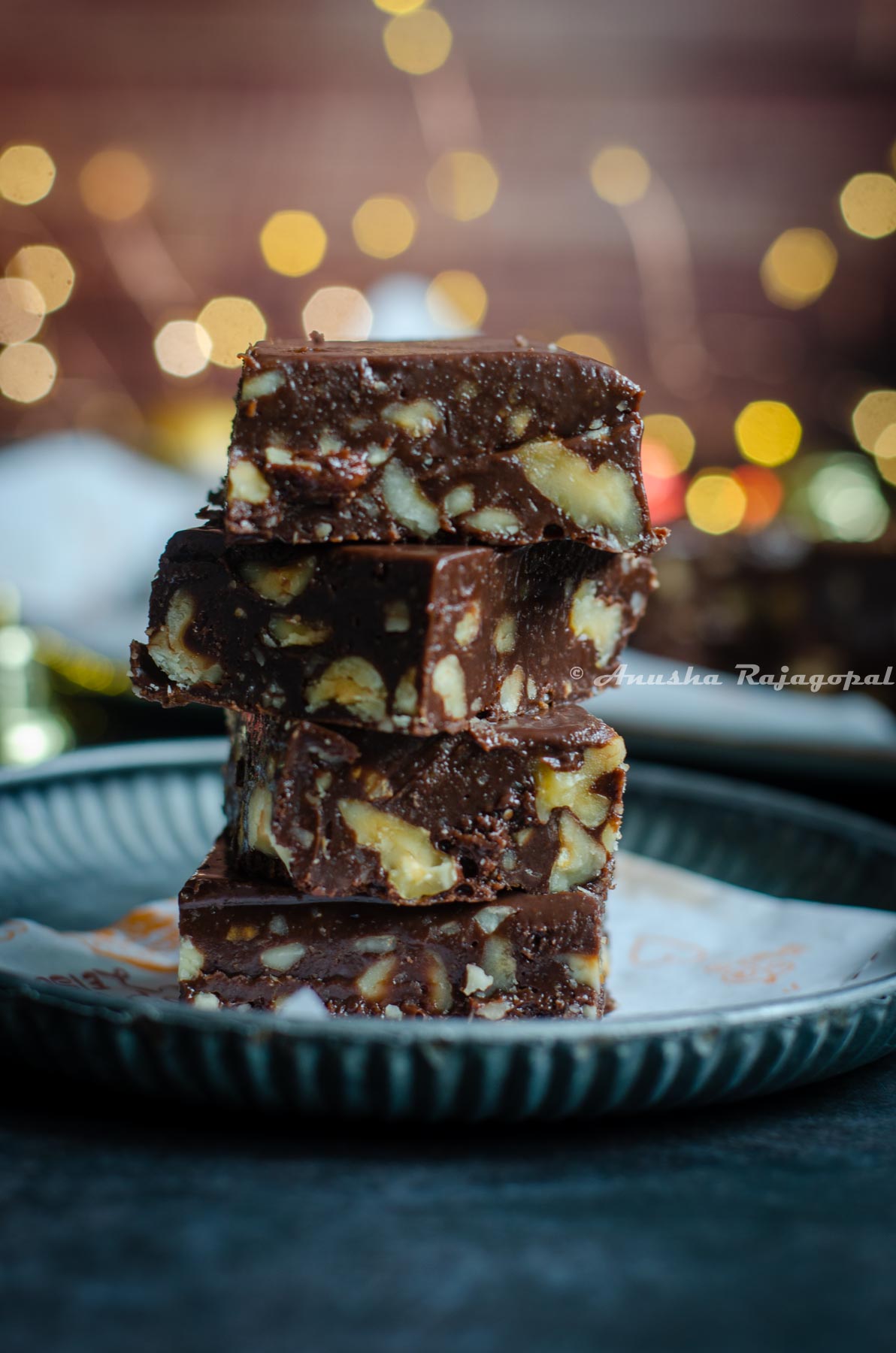 chocolate fudge stacked over a piece of parchment paper placed on a rustic metal tray. Christmas ornaments placed at the back. Fairy lights not in focus.