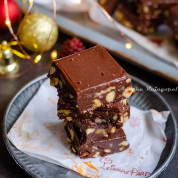 chocolate fudge stacked over a piece of parchment paper placed on a rustic metal tray. Christmas ornaments placed at the back. Fairy lights not in focus.