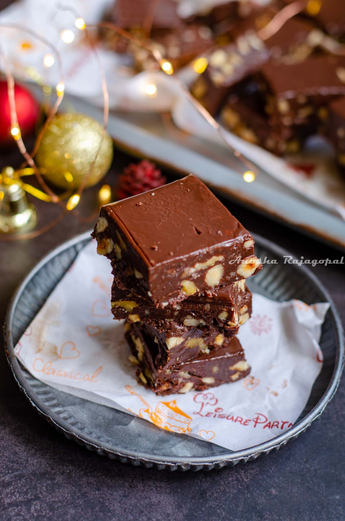 chocolate fudge stacked over a piece of parchment paper placed on a rustic metal tray. Christmas ornaments placed at the back. Fairy lights not in focus.