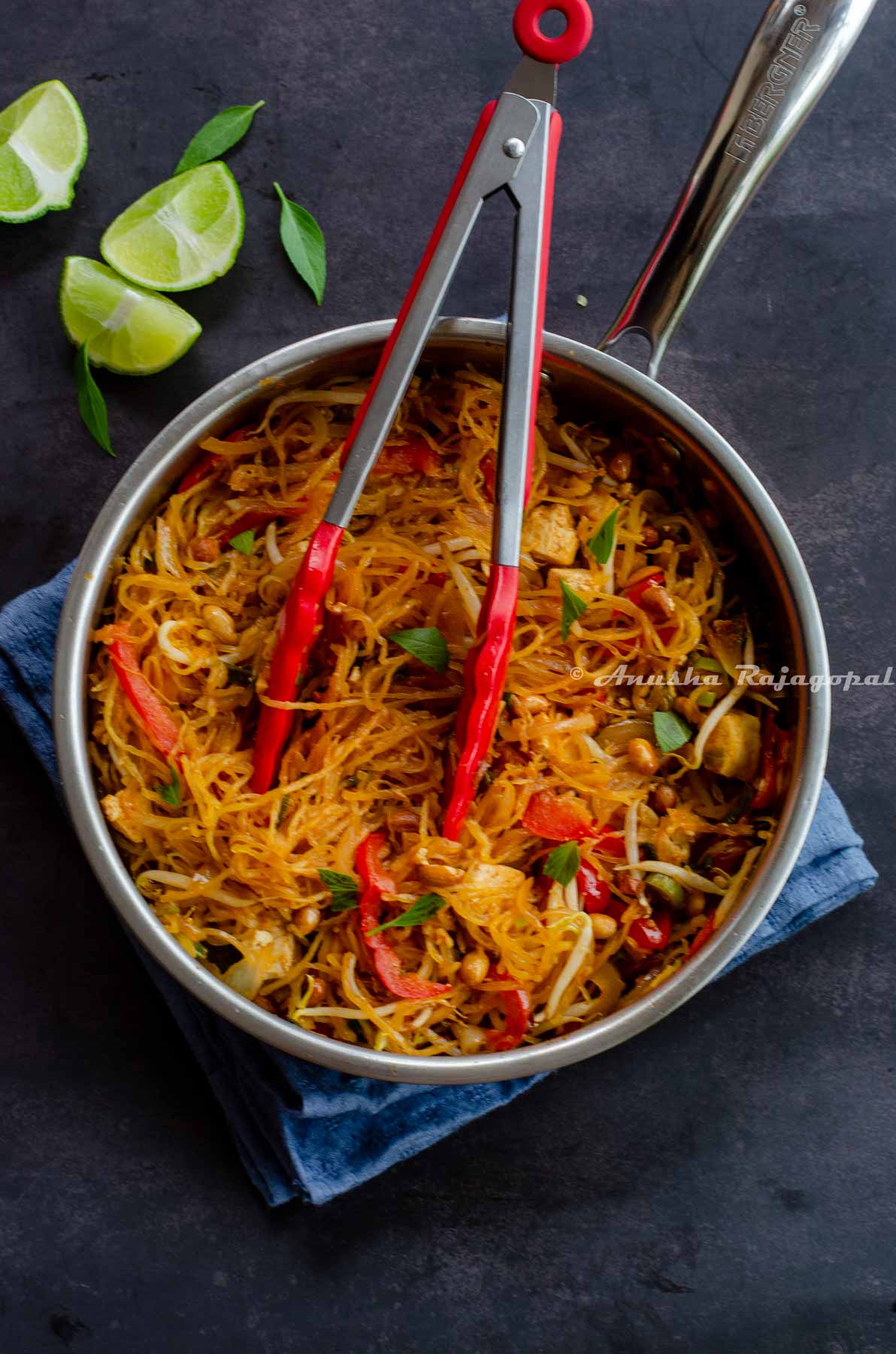 Spaghetti Squash Pad Thai in a saute pan with a pair of red tongs placed on the Pad Thai. Lime wedges and Thai basil at the background.