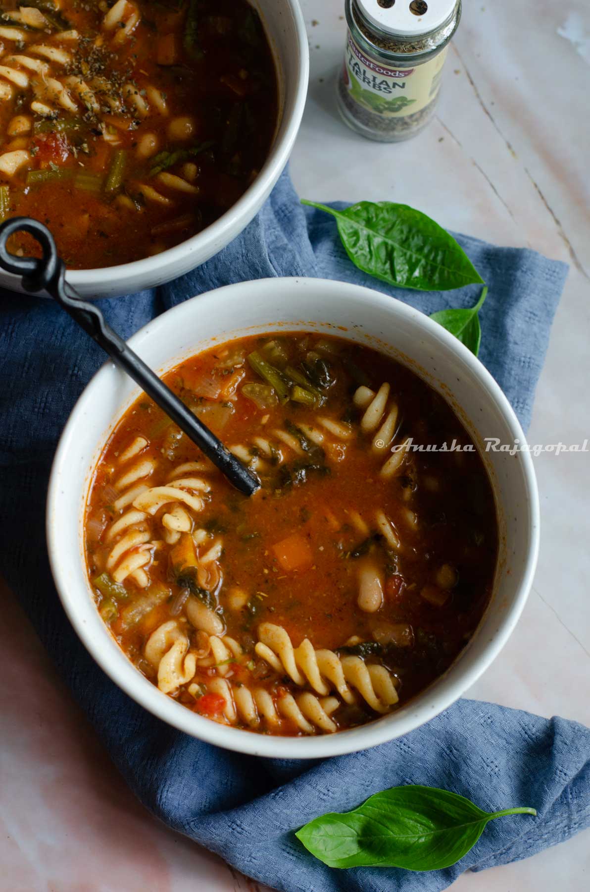 vegan minestrone soup serveVegan minestrone soup in a white bowl with a black handled spoon in it. The bowl is placed on a blue linen cloth. Fresh sweet basil leaves strewn around the bowl.