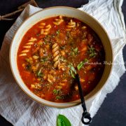 vegan minestrone soup served in a brown rimmed beige bowl with a black handled spoon in it. The bowl is placed on a white linen cloth. Fresh sweet basil leaves strewn around the bowl.