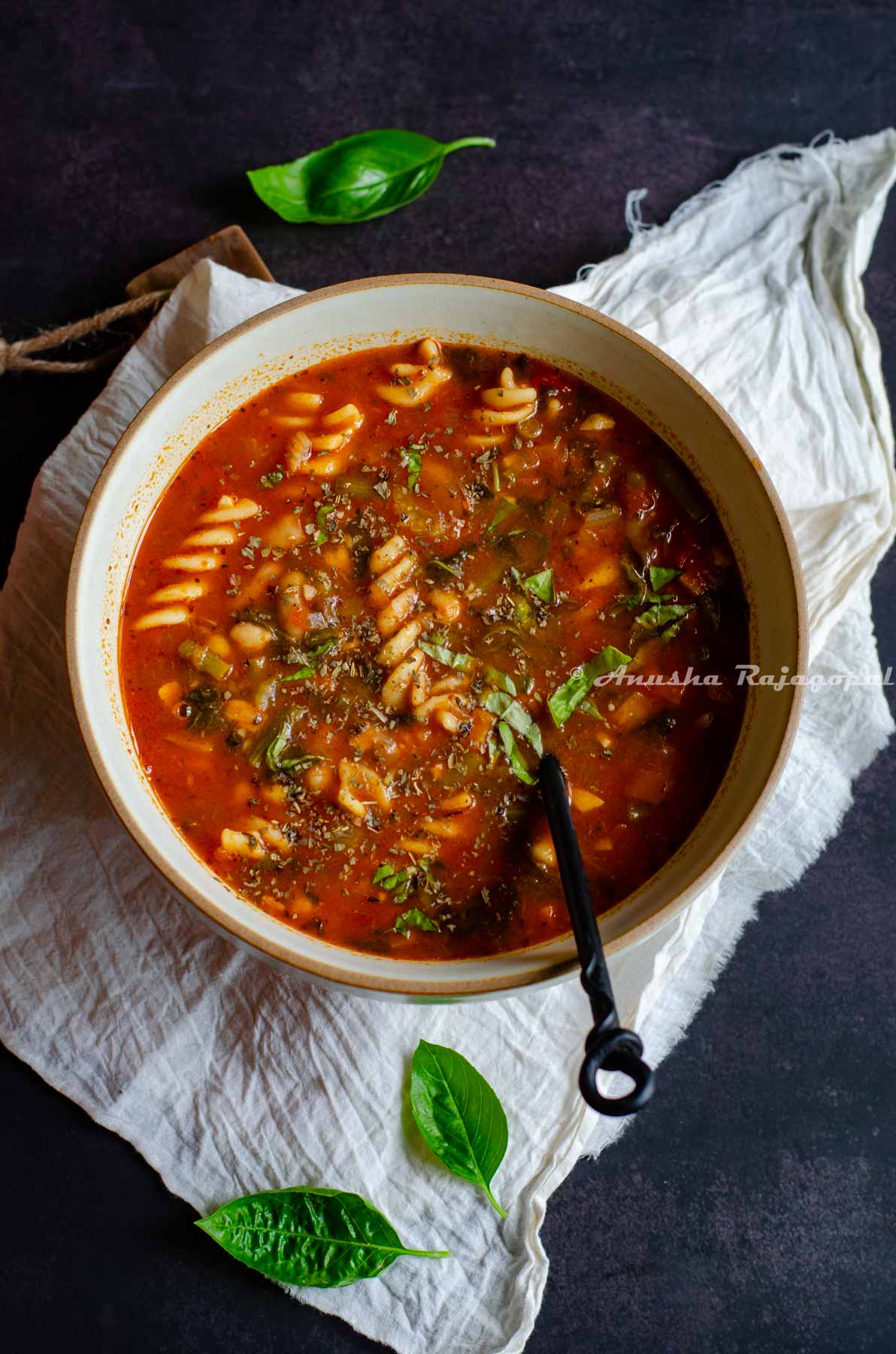 vegan minestrone soup served in a brown rimmed beige bowl with a black handled spoon in it. The bowl is placed on a white linen cloth. Fresh sweet basil leaves strewn around the bowl.