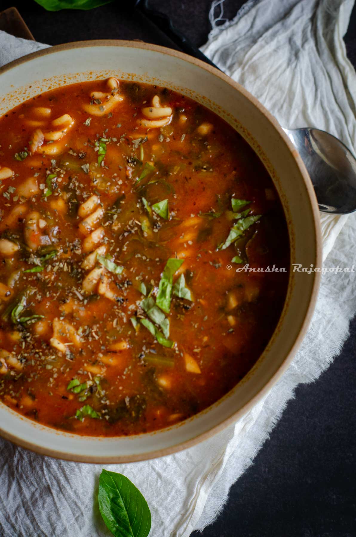 vegan minestrone soup served in a brown rimmed beige bowl with a black handled spoon in it. The bowl is placed on a white linen cloth. Fresh sweet basil leaves strewn around the bowl.