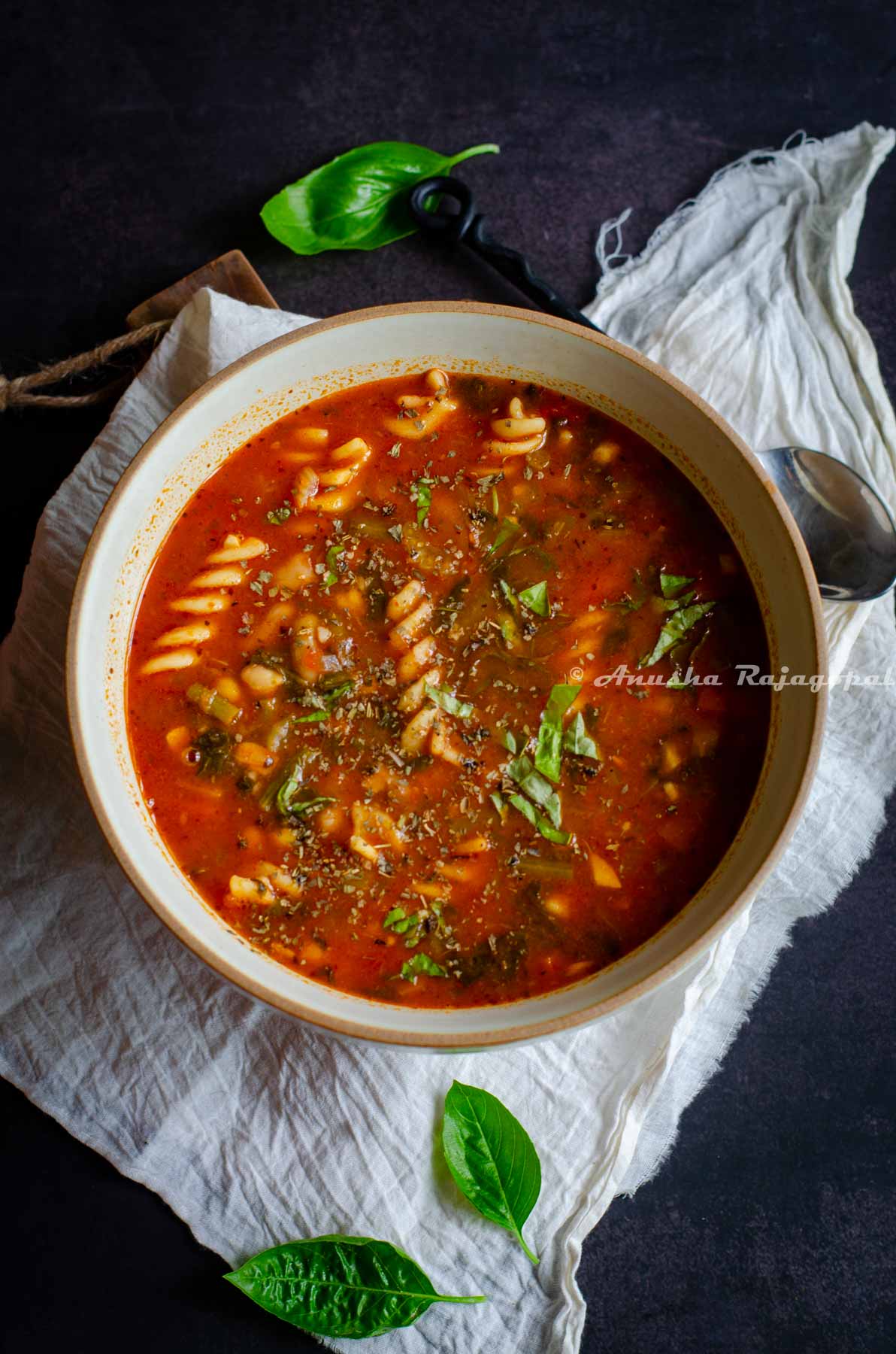 vegan minestrone soup served in a brown rimmed beige bowl .The bowl is placed on a white linen cloth. Fresh sweet basil leaves strewn around the bowl.