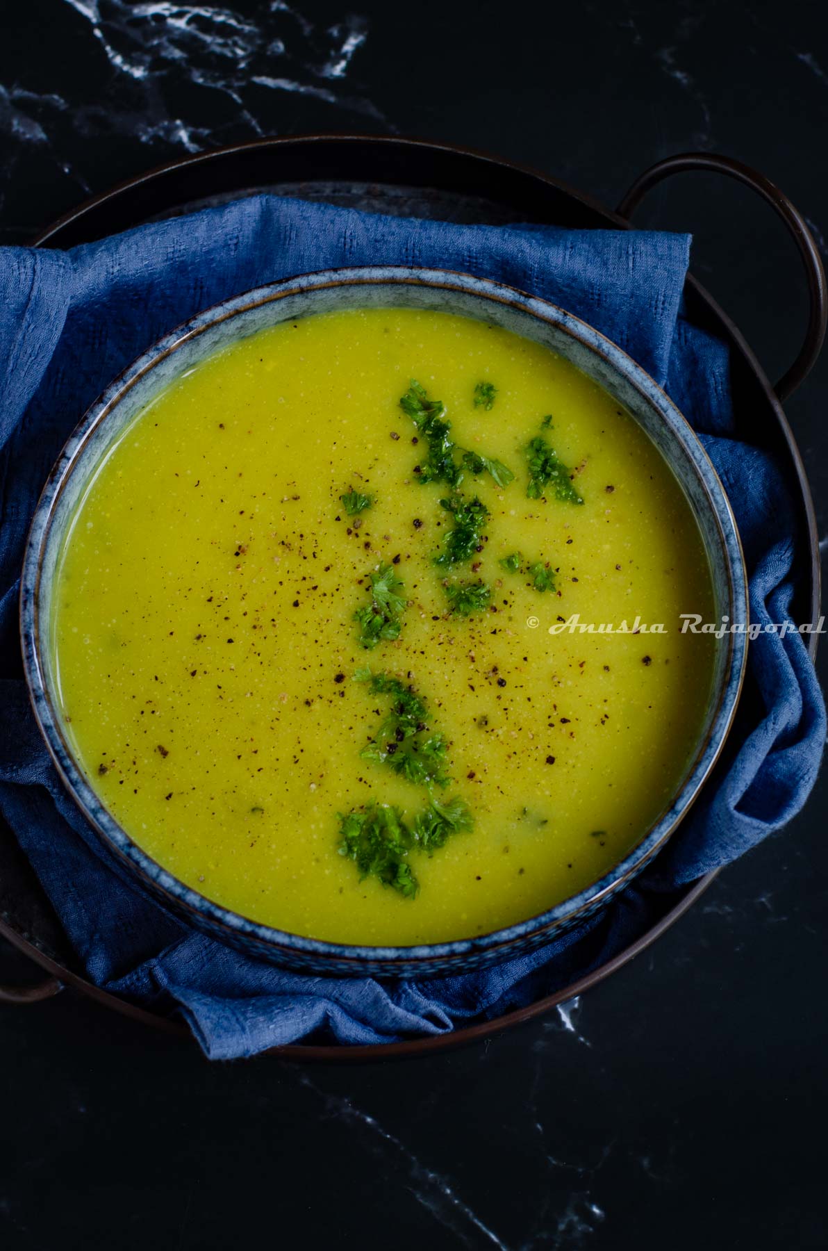 Instant Pot cauliflower soup served in a greyish black shallow bowl placed on a rustic metal tray with a blue linen cloth beneath it.