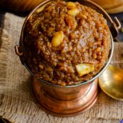 Sakkarai Pongal, a traditional Tamil style dessert served in a copper serving bucket. Cashews on the top as garnish. The serving dish has been placed on a burlap mat. Brass spoons and wooden bowls of rice and lentil are at the background.