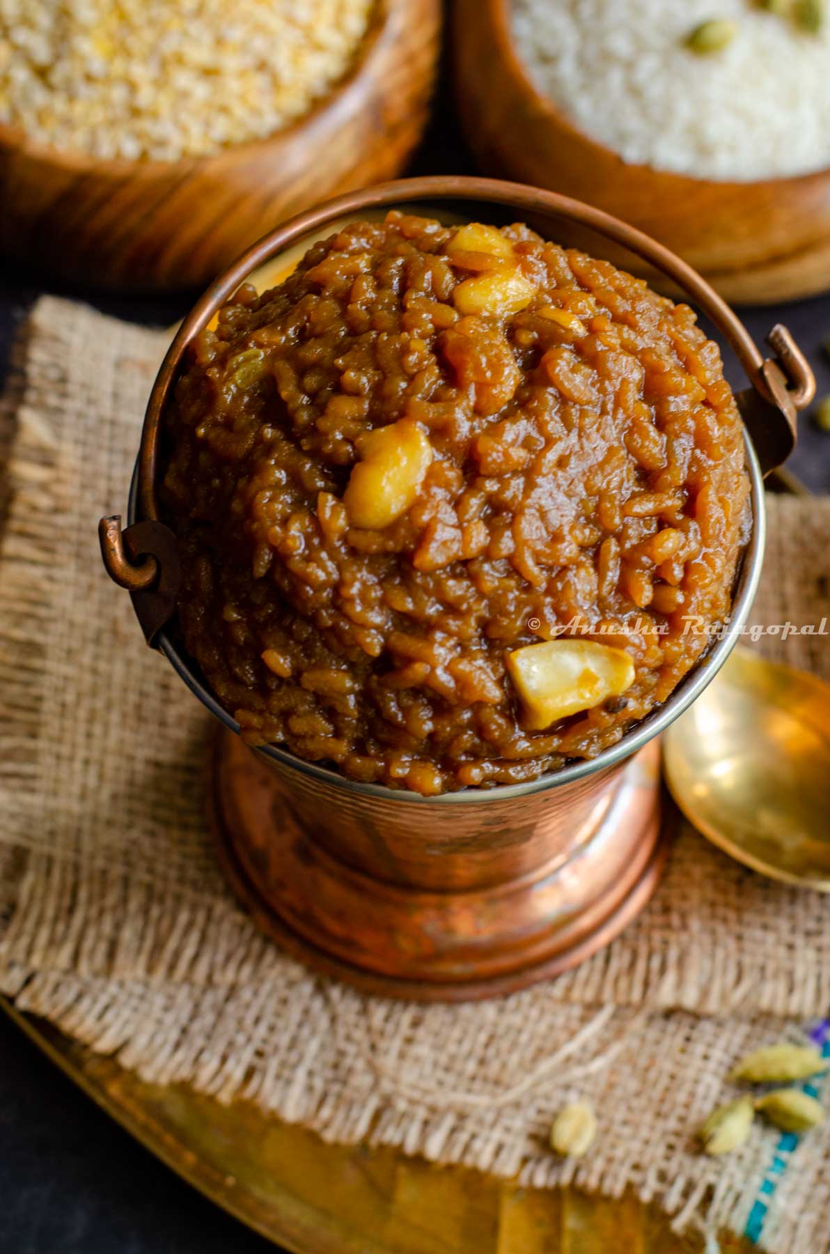 Sakkarai Pongal, a traditional Tamil style dessert served in a copper serving bucket. Cashews on the top as garnish. The serving dish has been placed on a burlap mat. Brass spoons and wooden bowls of rice and lentil are at the background.