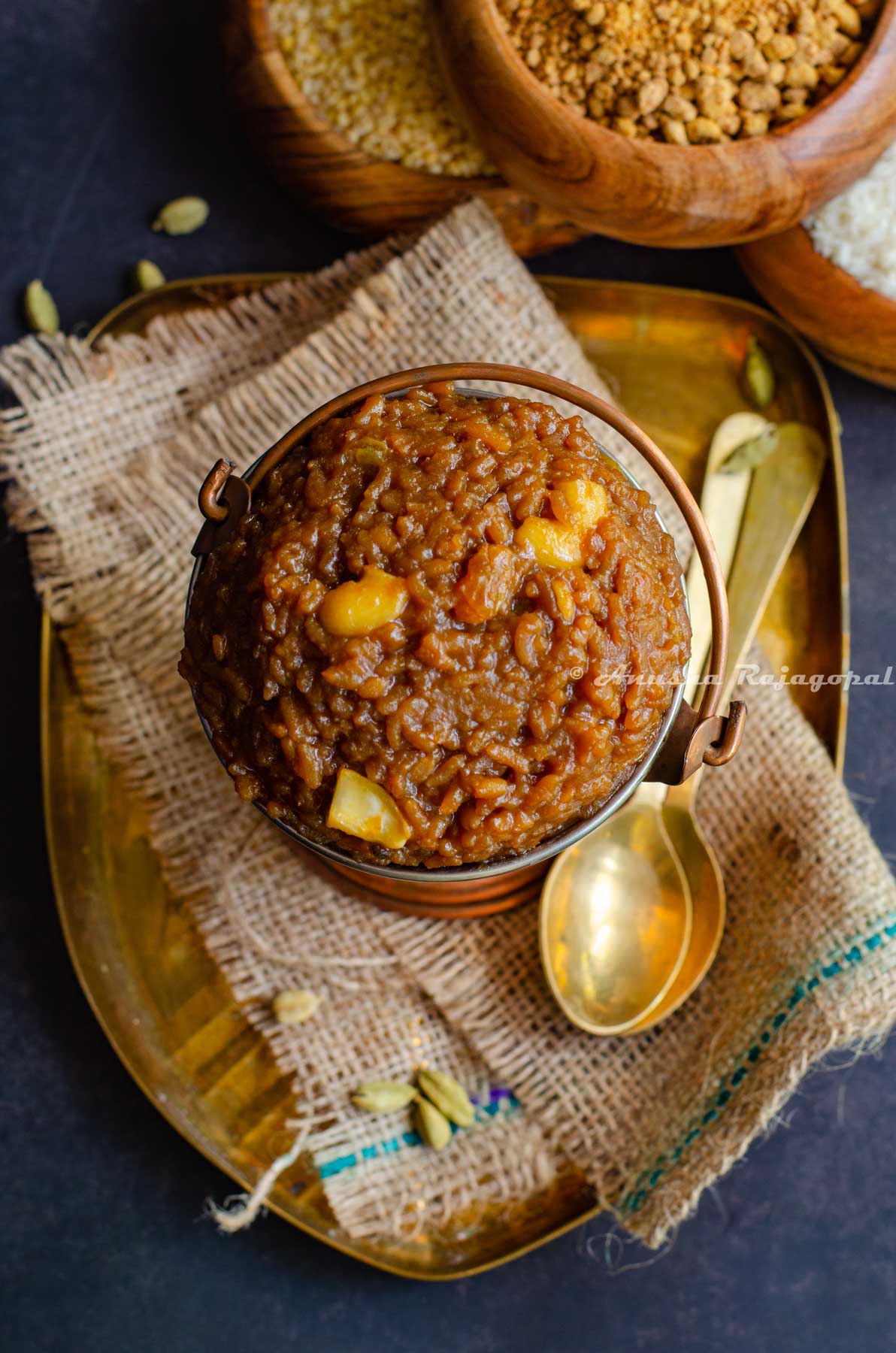 Sakkarai Pongal, a traditional Tamil style dessert served in a copper serving bucket. Cashews on the top as garnish. The serving dish has been placed on a burlap mat. Brass spoons and wooden bowls of rice and lentil are at the background.