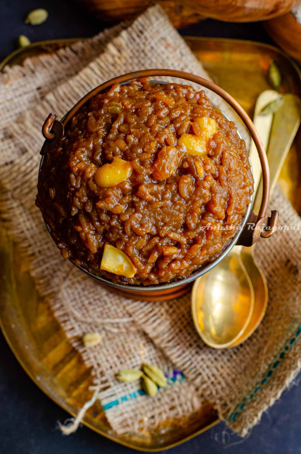 Sakkarai Pongal, a traditional Tamil style dessert served in a copper serving bucket. Cashews on the top as garnish. The serving dish has been placed on a burlap mat. Brass spoons and wooden bowls of rice and lentil are at the background.