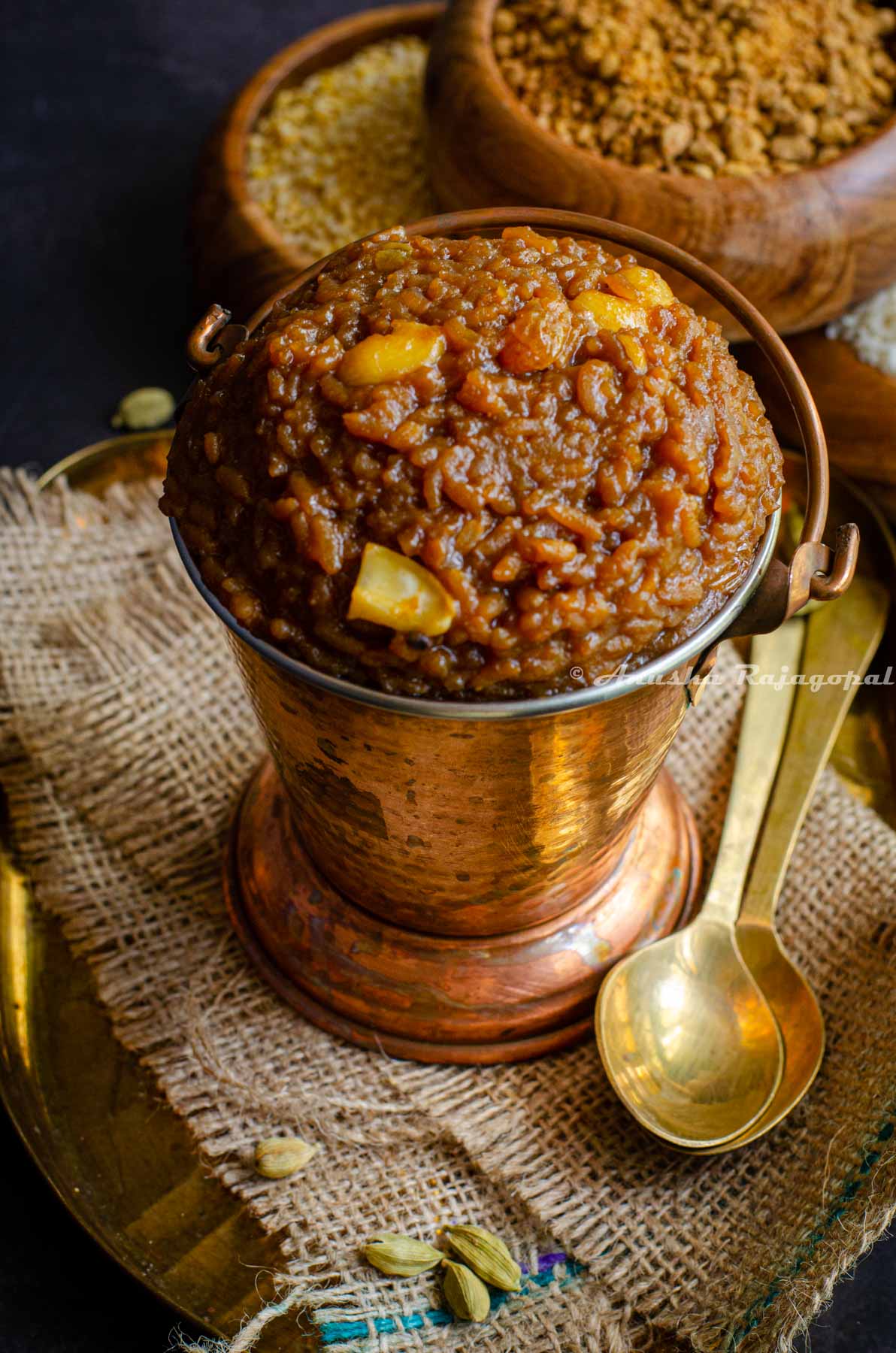 Sakkarai Pongal, a traditional Tamil style dessert served in a copper serving bucket. Cashews on the top as garnish. The serving dish has been placed on a burlap mat. Brass spoons and wooden bowls of rice and lentil are at the background.