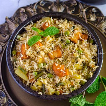 Brinji rice served in a black bowl placed over a rustic brown platter. Mint leaves by the side and chilled onion raita at the background.