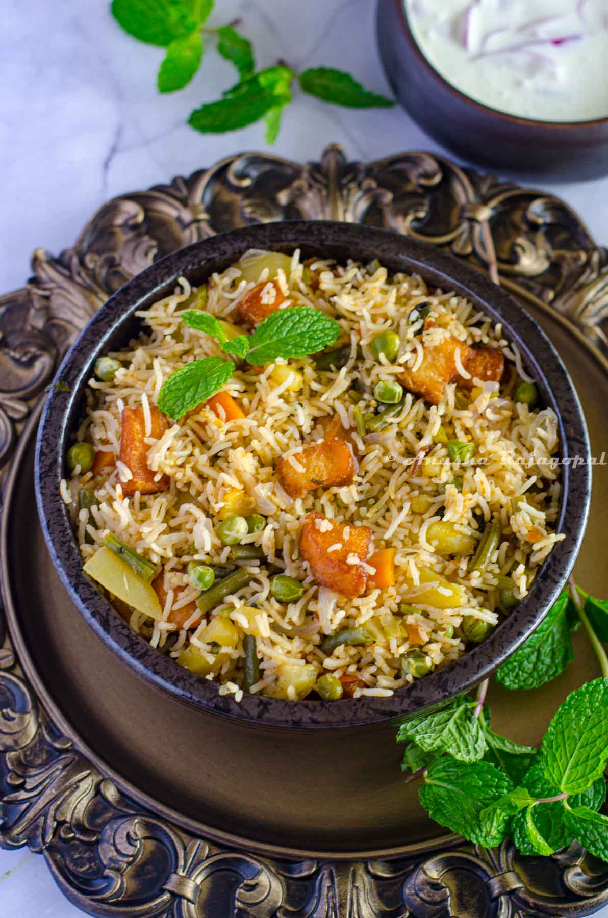Brinji rice served in a black bowl placed over a rustic brown platter. Mint leaves by the side and chilled onion raita at the background.