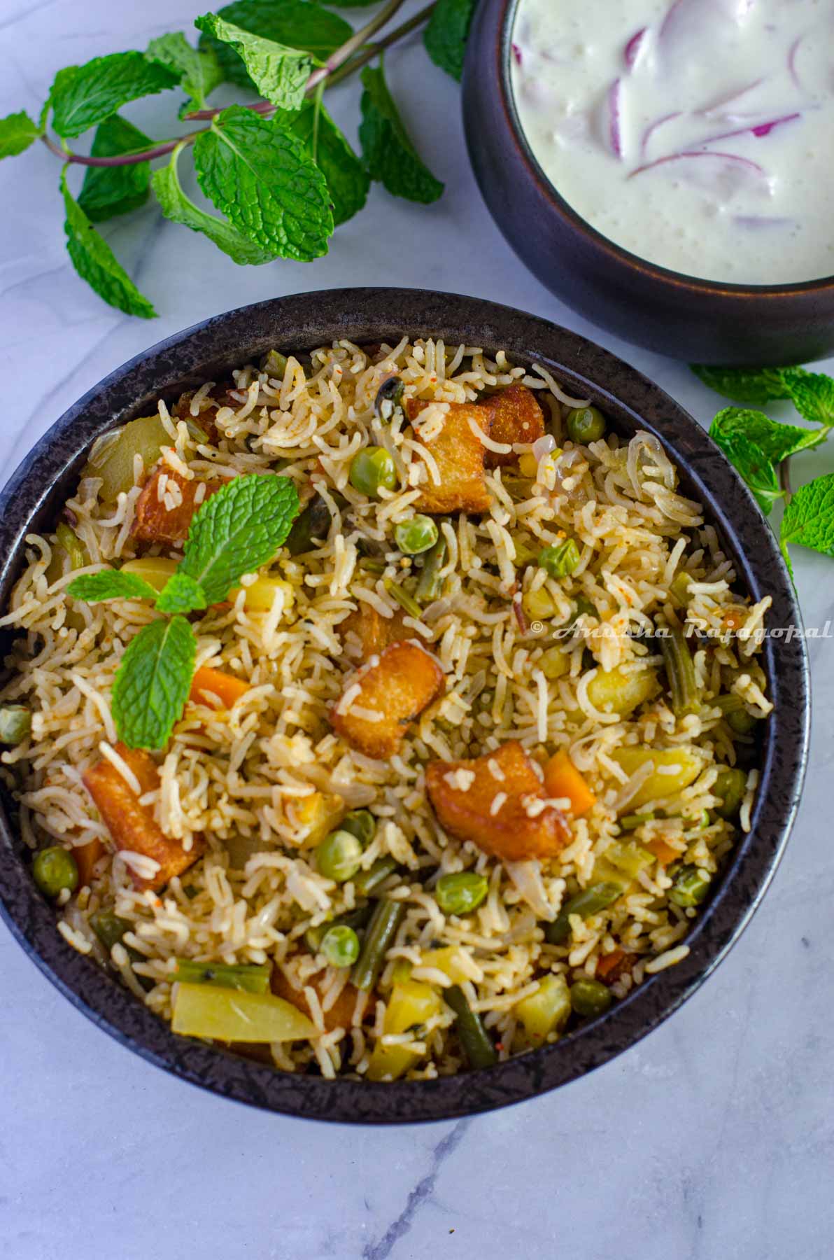 Brinji rice served in a black bowl. Mint leaves by the side and chilled onion raita at the background.